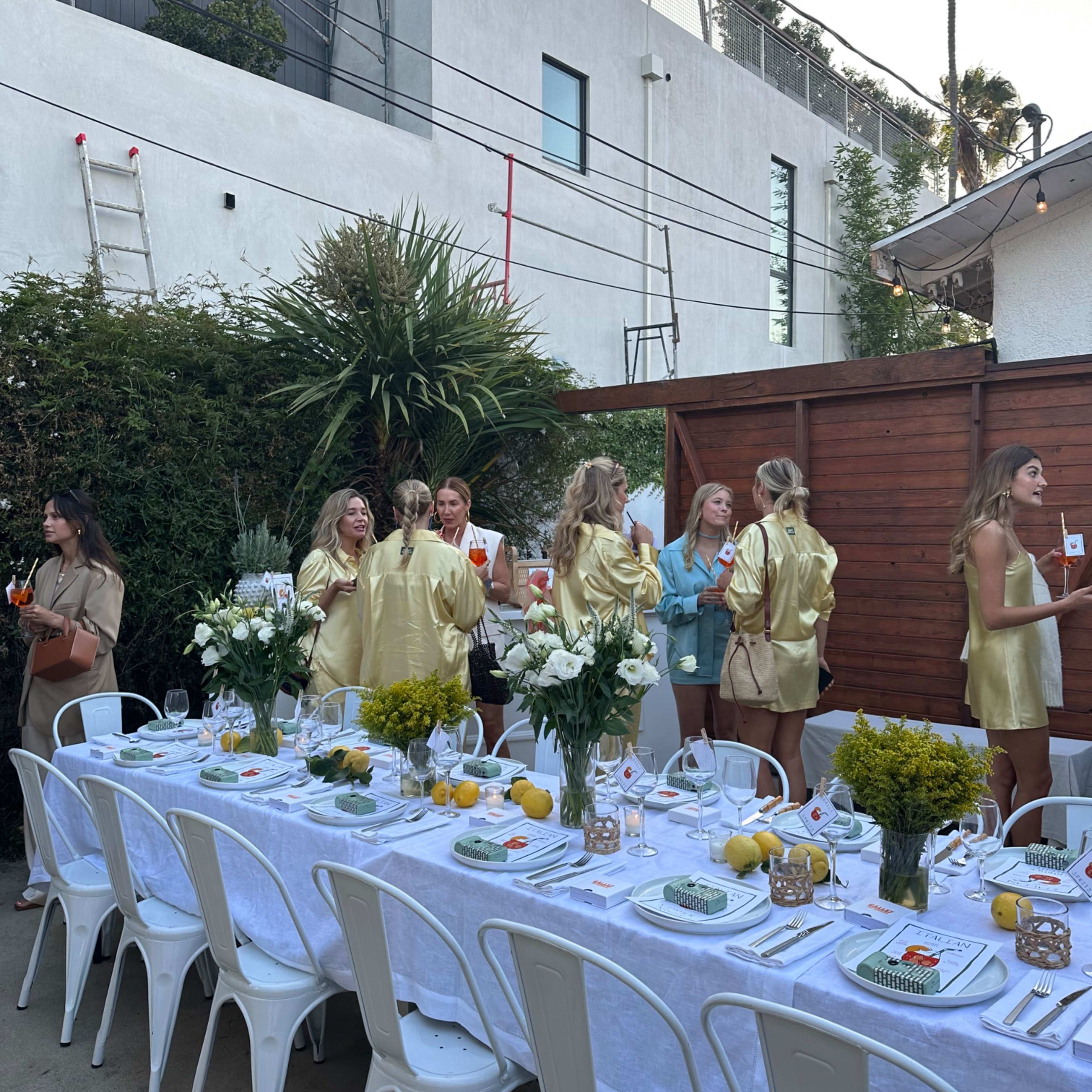 A long dining table is set outdoors with white linens and centerpieces, while a group of women in golden outfits socialize nearby.