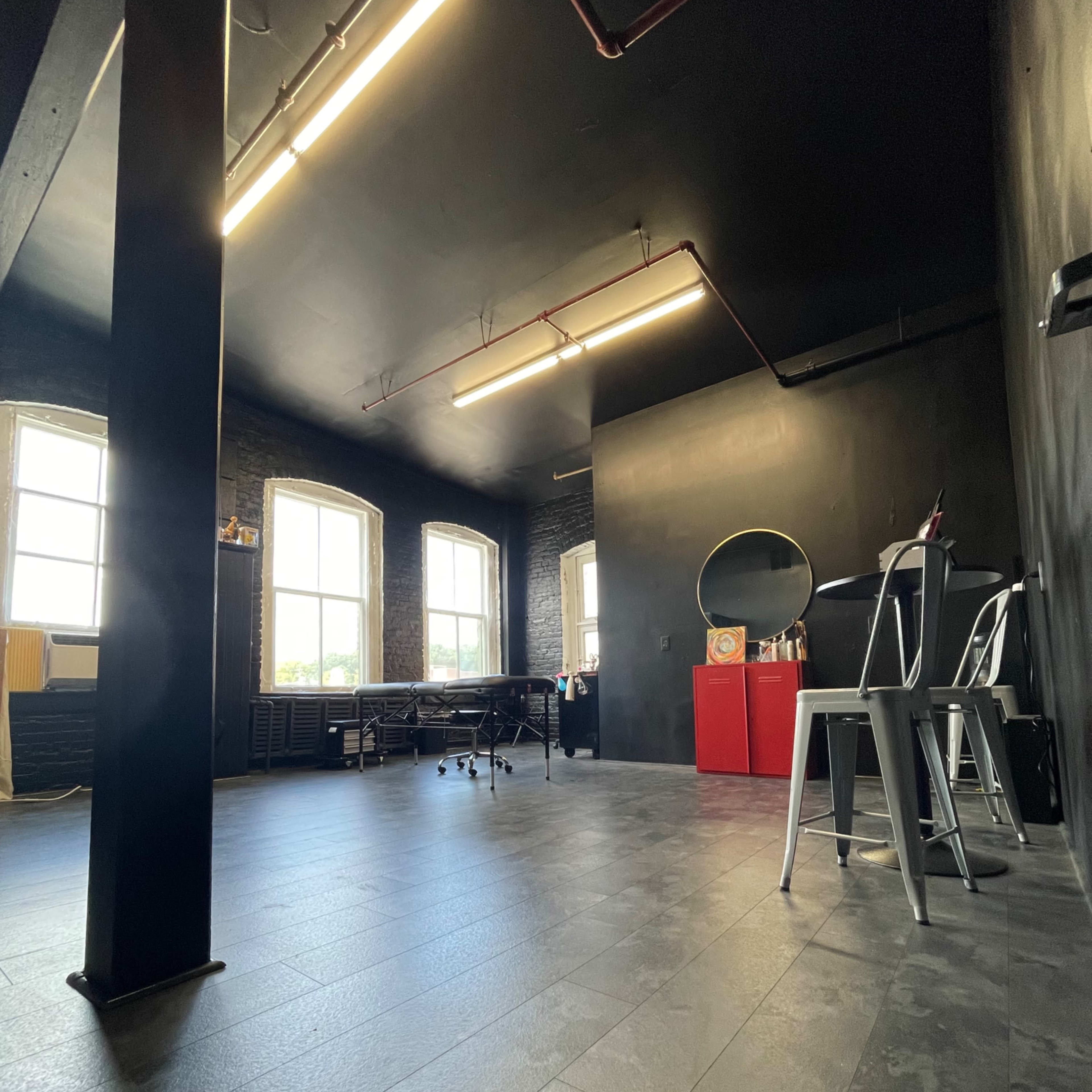 A spacious studio with large windows, a black ceiling and walls, a red storage unit, a mirror, and metal chairs arranged around a table.
