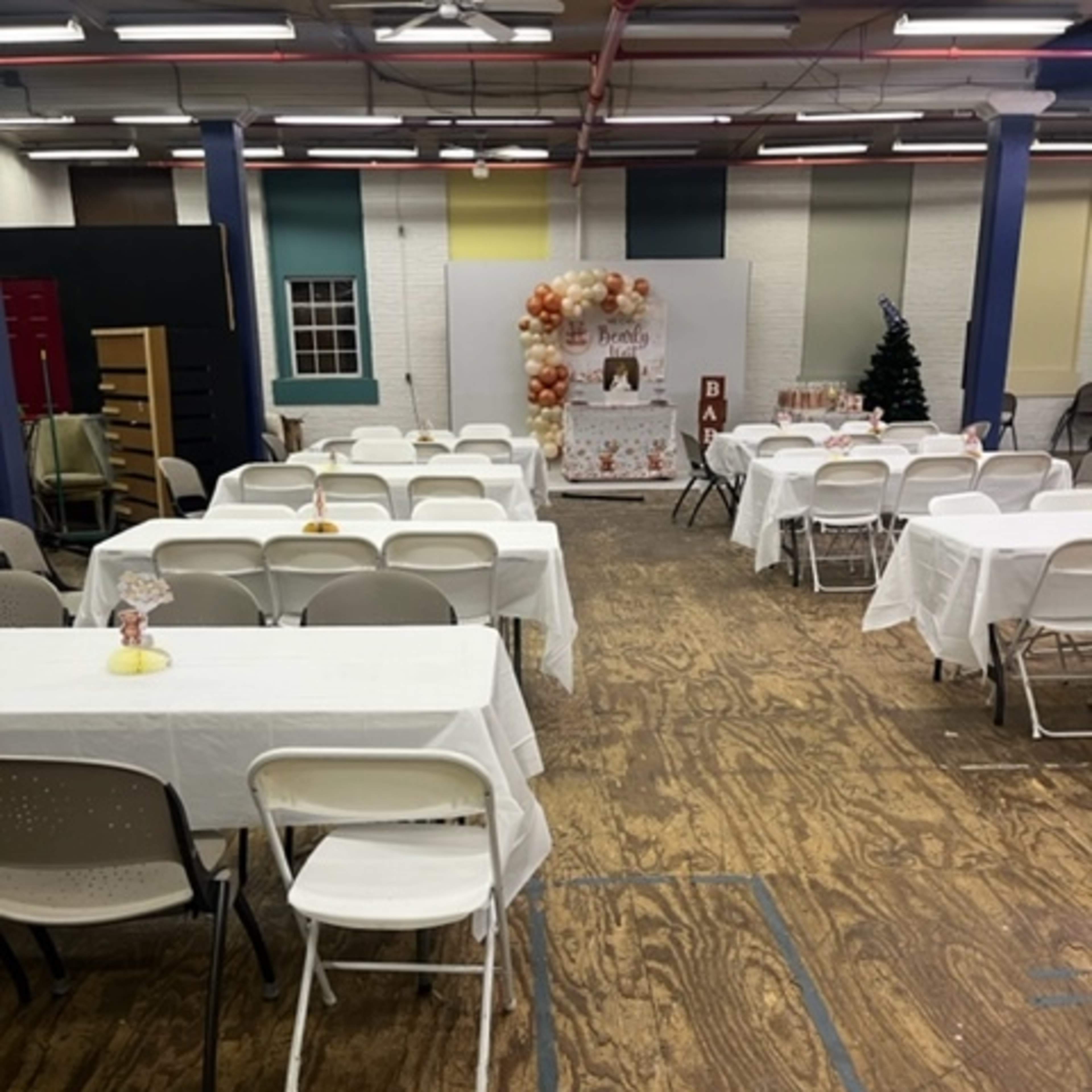 An indoor event space with several tables covered in white tablecloths, set up for an occasion, and festive decorations in the background.