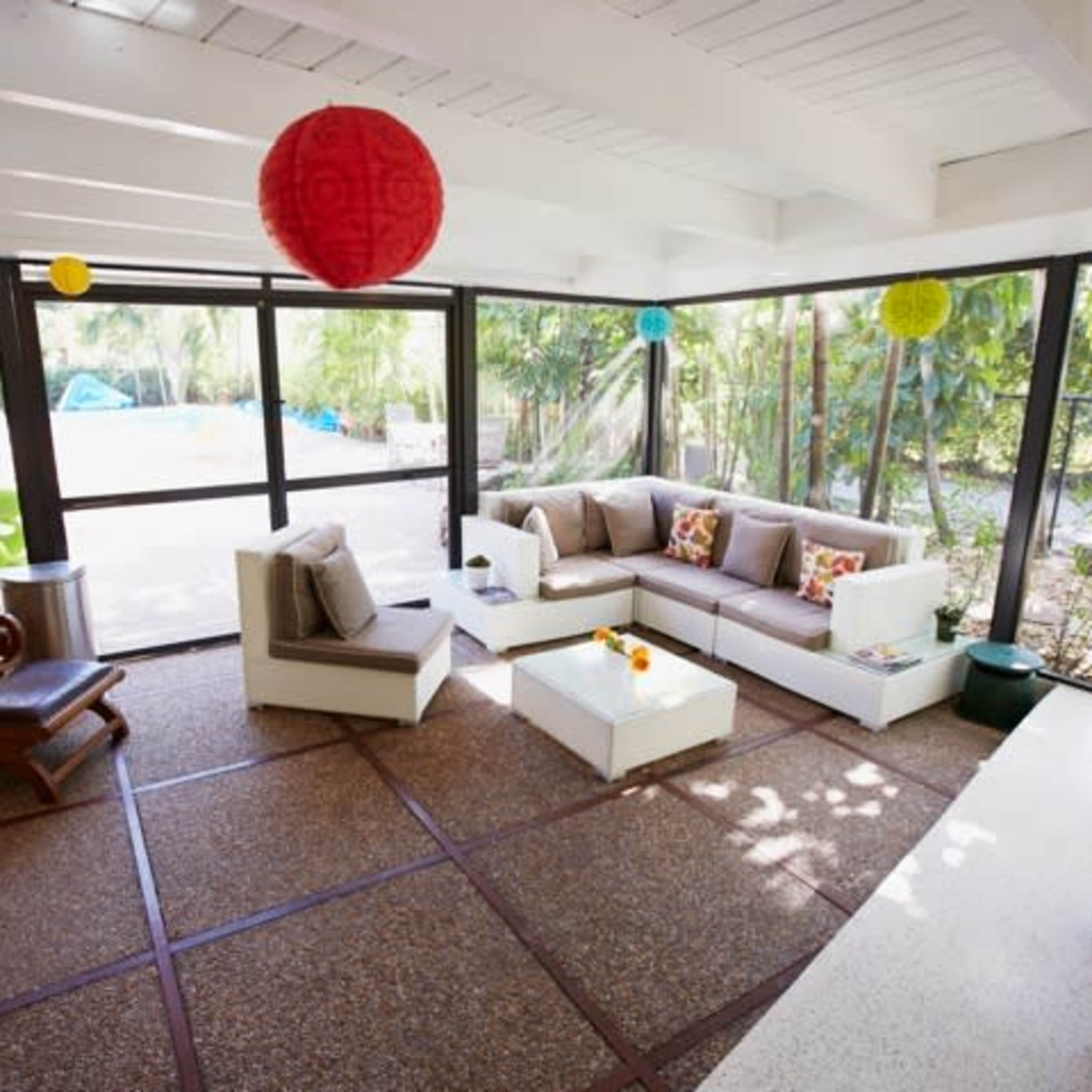 A bright, enclosed living area with a modern sectional sofa, a coffee table, and a rocking chair, surrounded by large windows overlooking a garden.