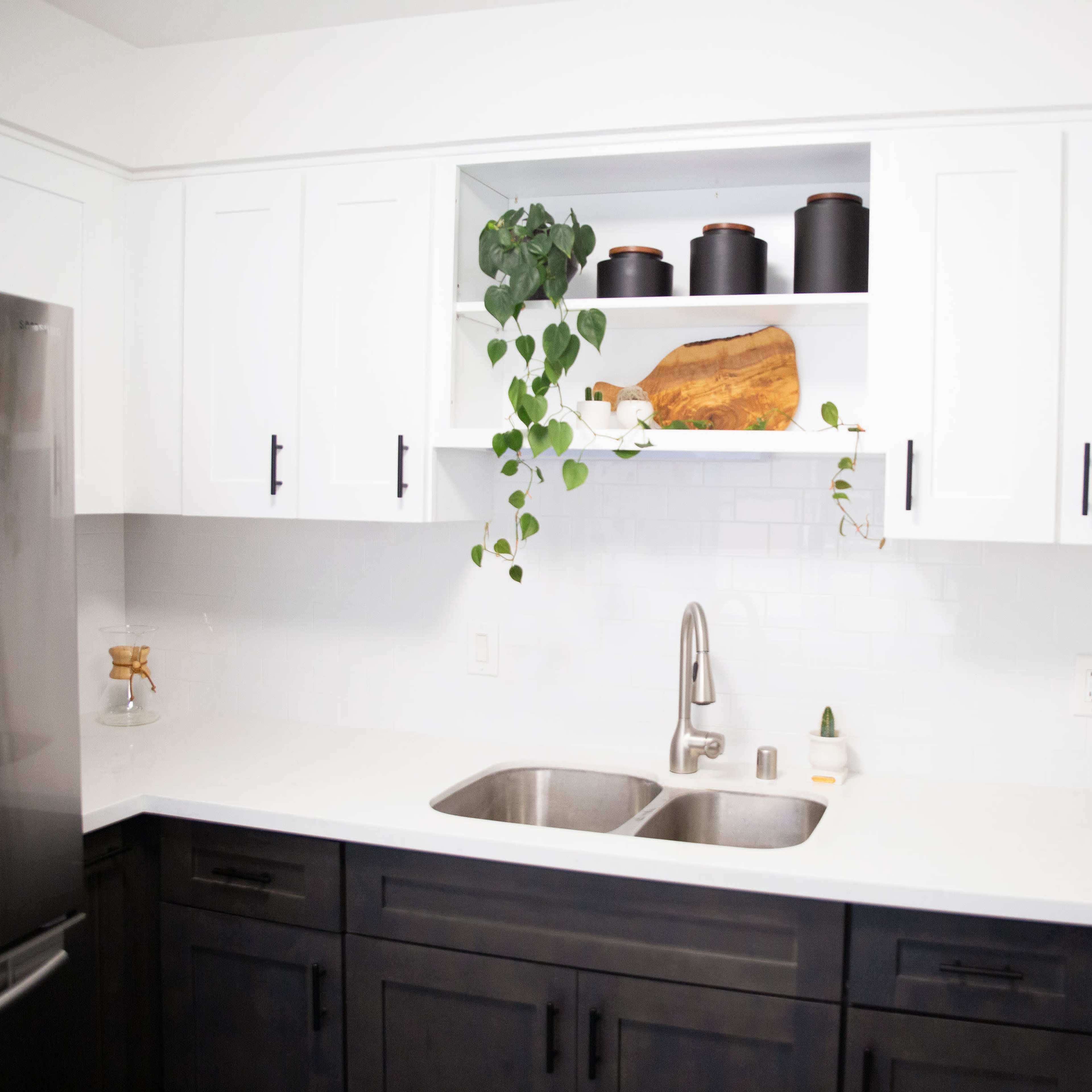 A modern kitchen with black lower cabinets, white upper cabinets, and a double sink underneath a wall-mounted shelf displaying decorative items and plants.