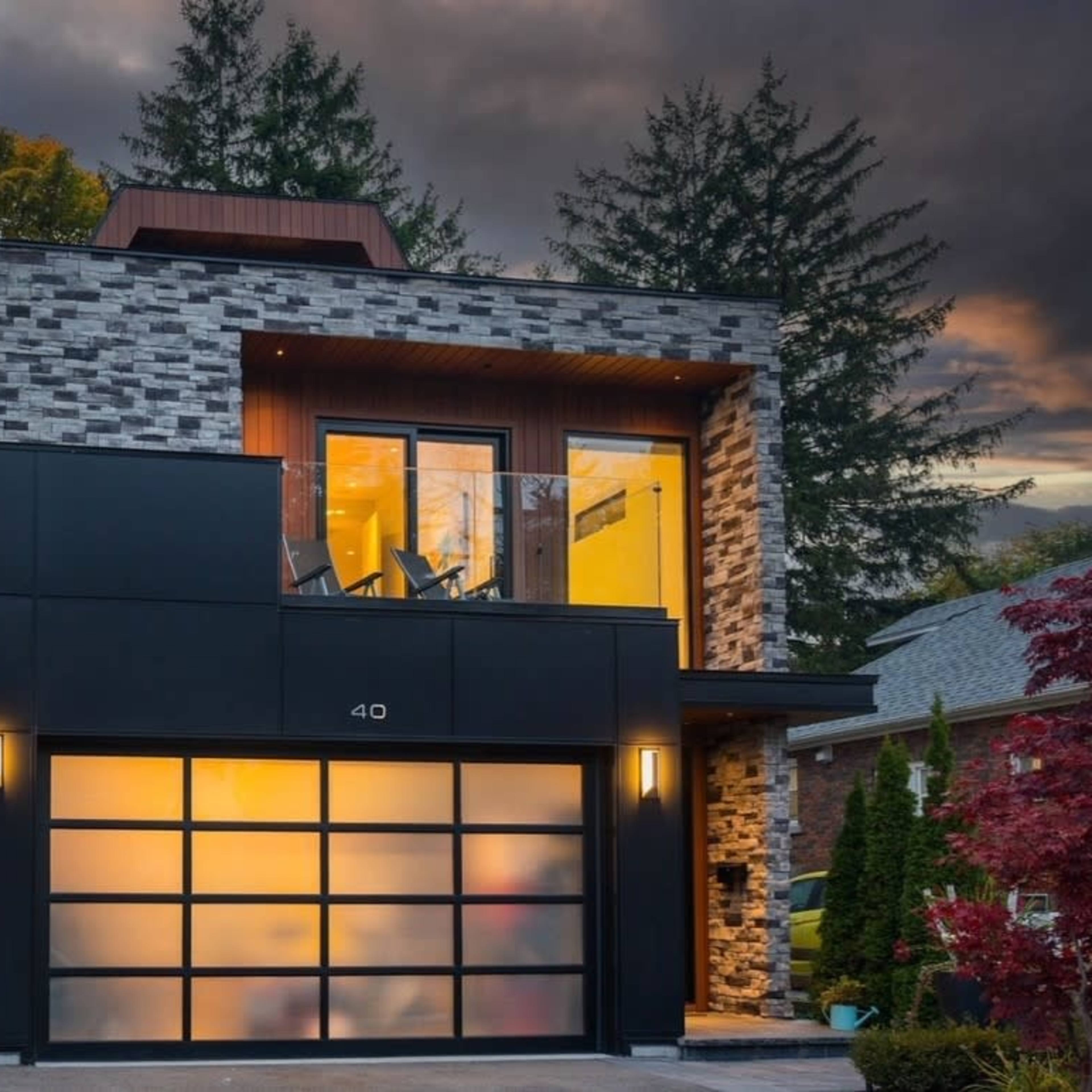 A modern two-story house with a stone and dark panel exterior, featuring a garage with frosted glass doors and a balcony on the upper floor.