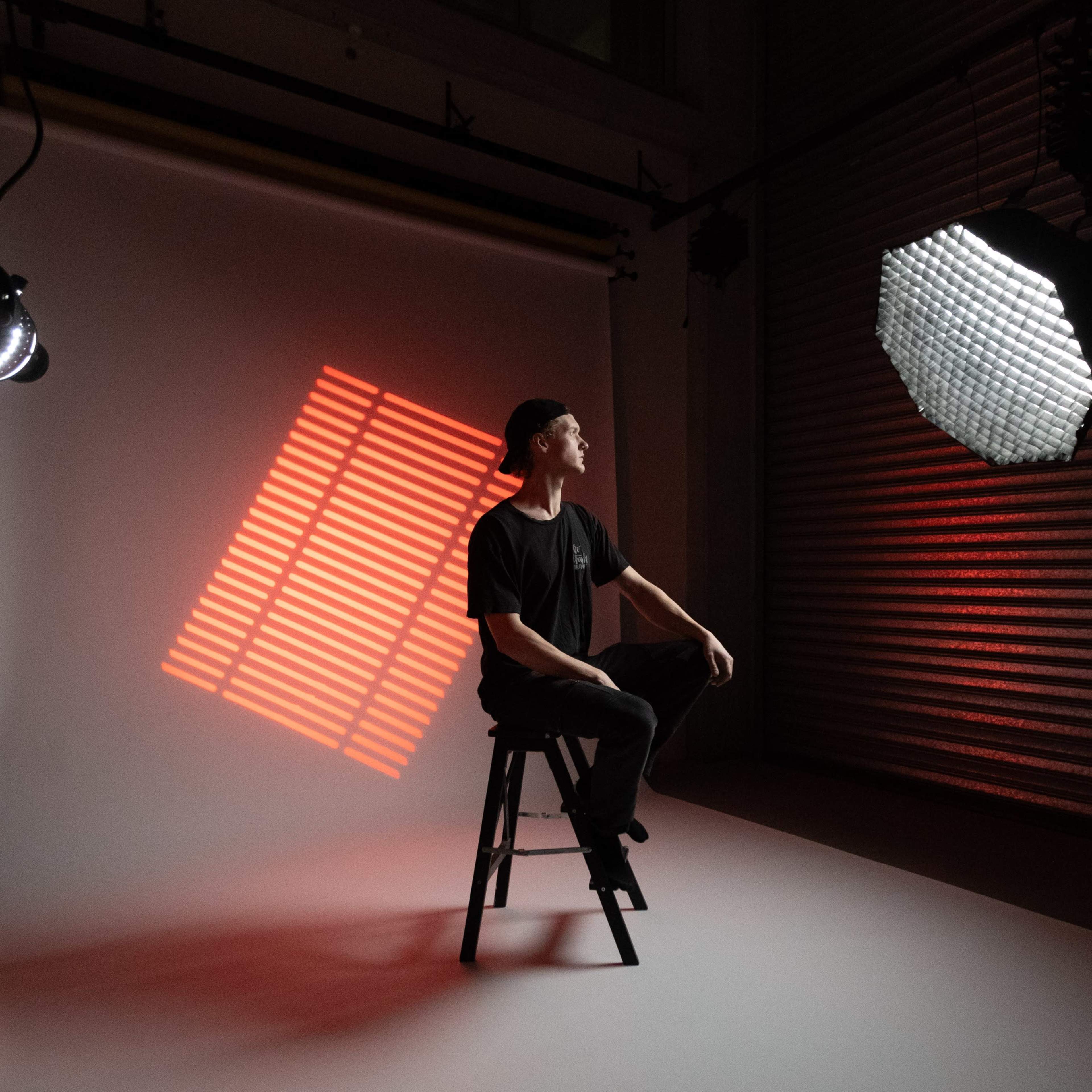 A person sits on a stool in a studio, with a shadow of striped light projected onto the backdrop.