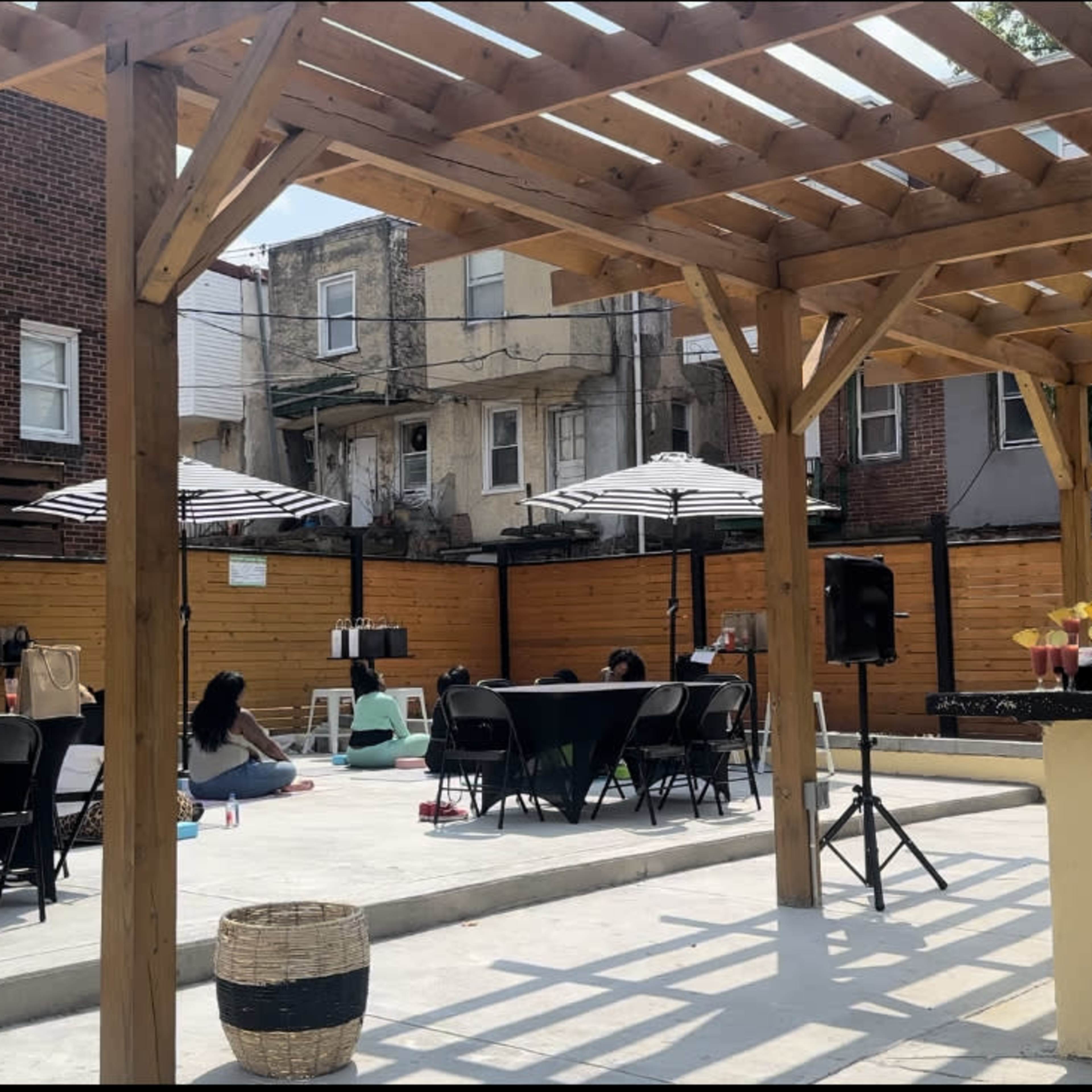 An outdoor patio area featuring several tables and chairs, a wooden pergola, and striped umbrellas, with people seated around the space.