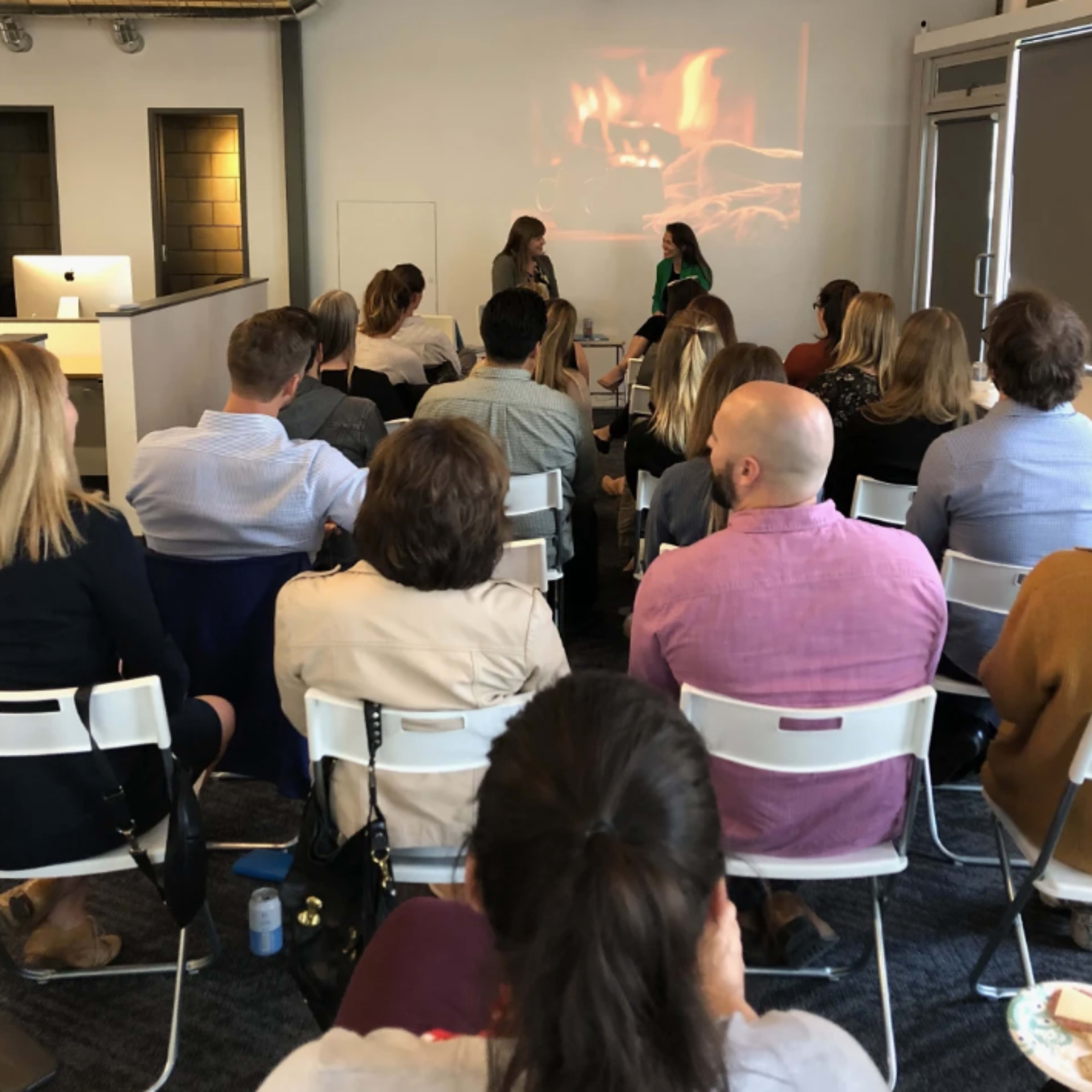 A group of people is seated in a conference room facing two women engaged in a discussion, with a projected image of a fireplace displayed behind them.