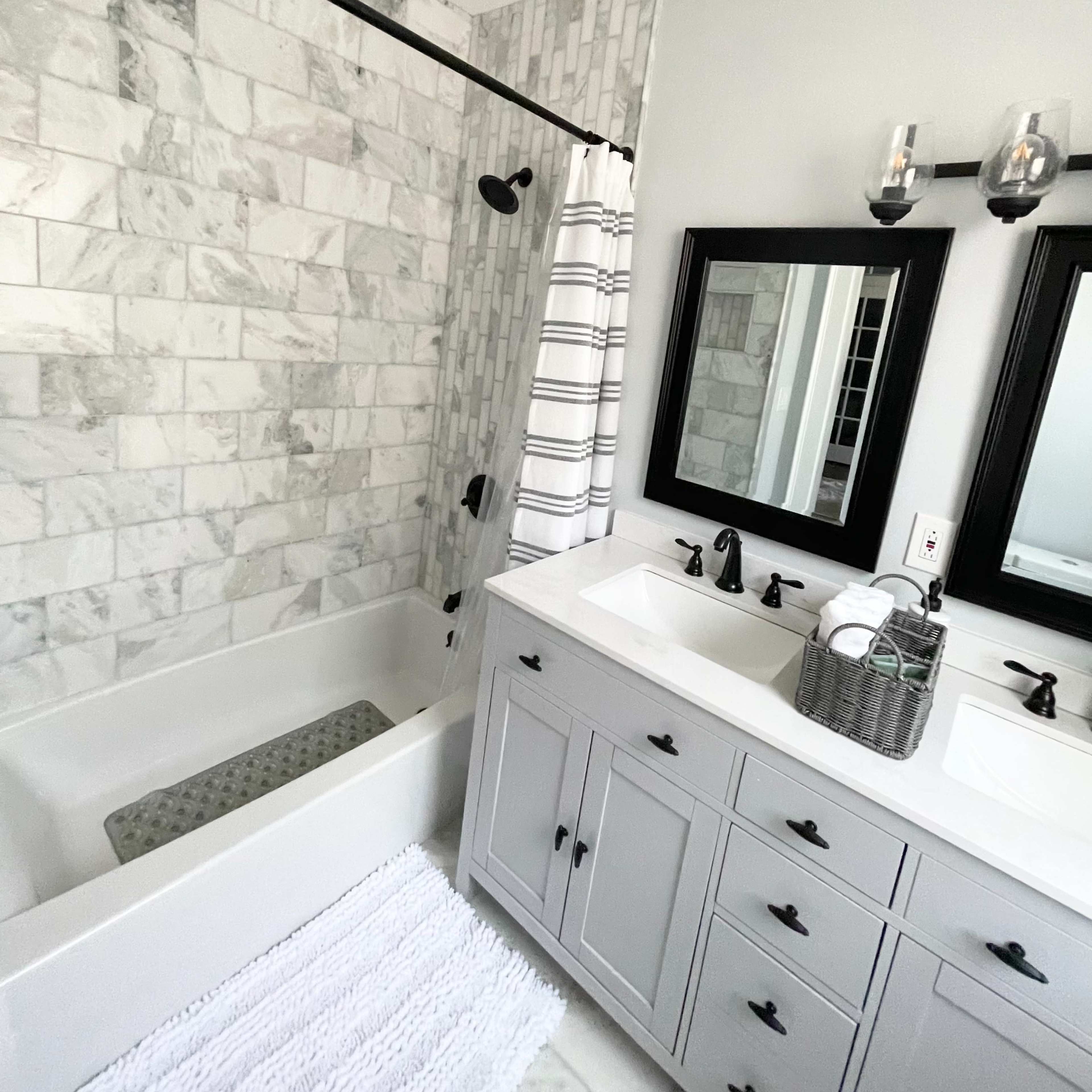 A modern bathroom featuring a marble-tiled shower area, a bathtub, dual sinks with a mirrored vanity, and a gray cabinetry.