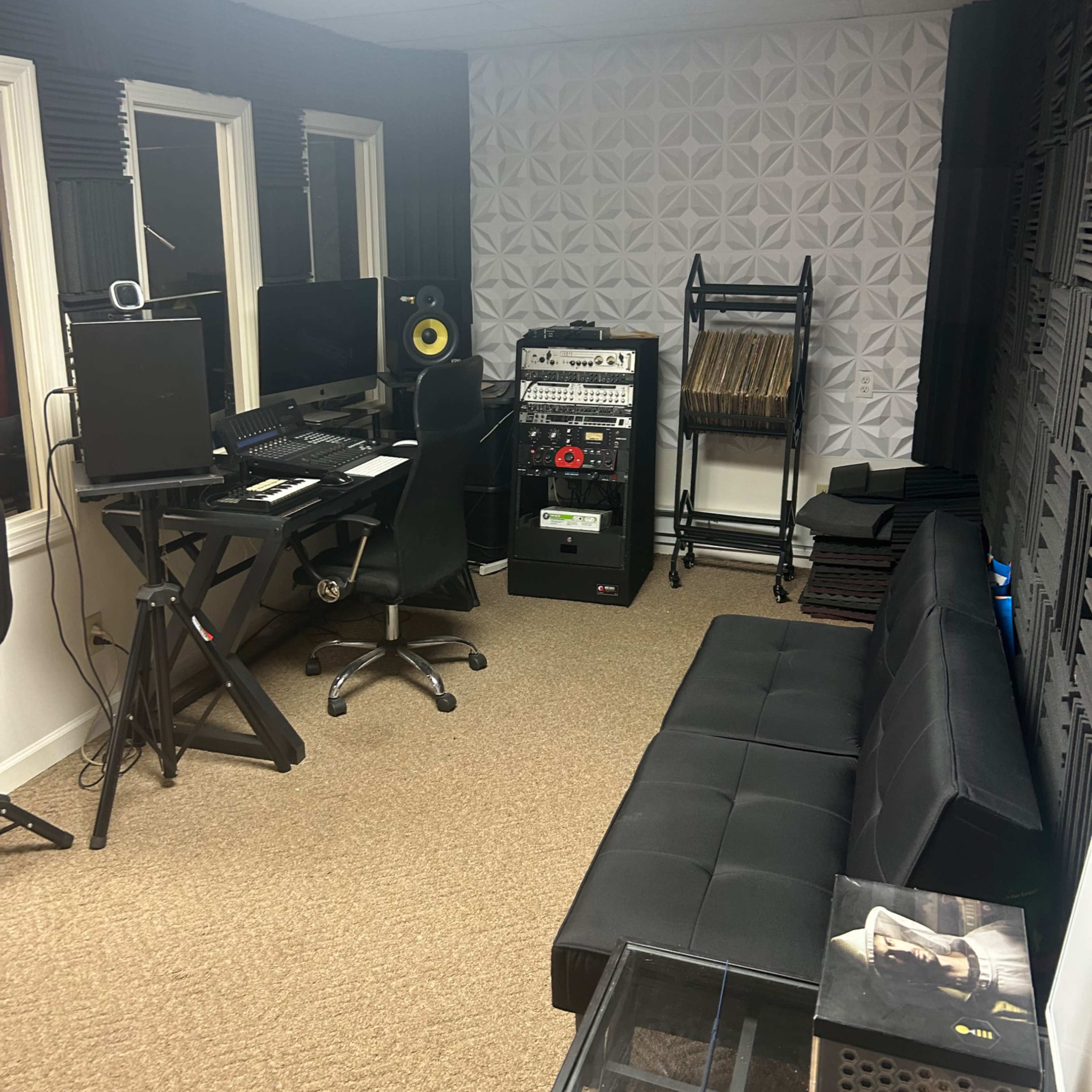 A small recording studio with a computer workstation, a black sofa, and audio equipment arranged against the walls.