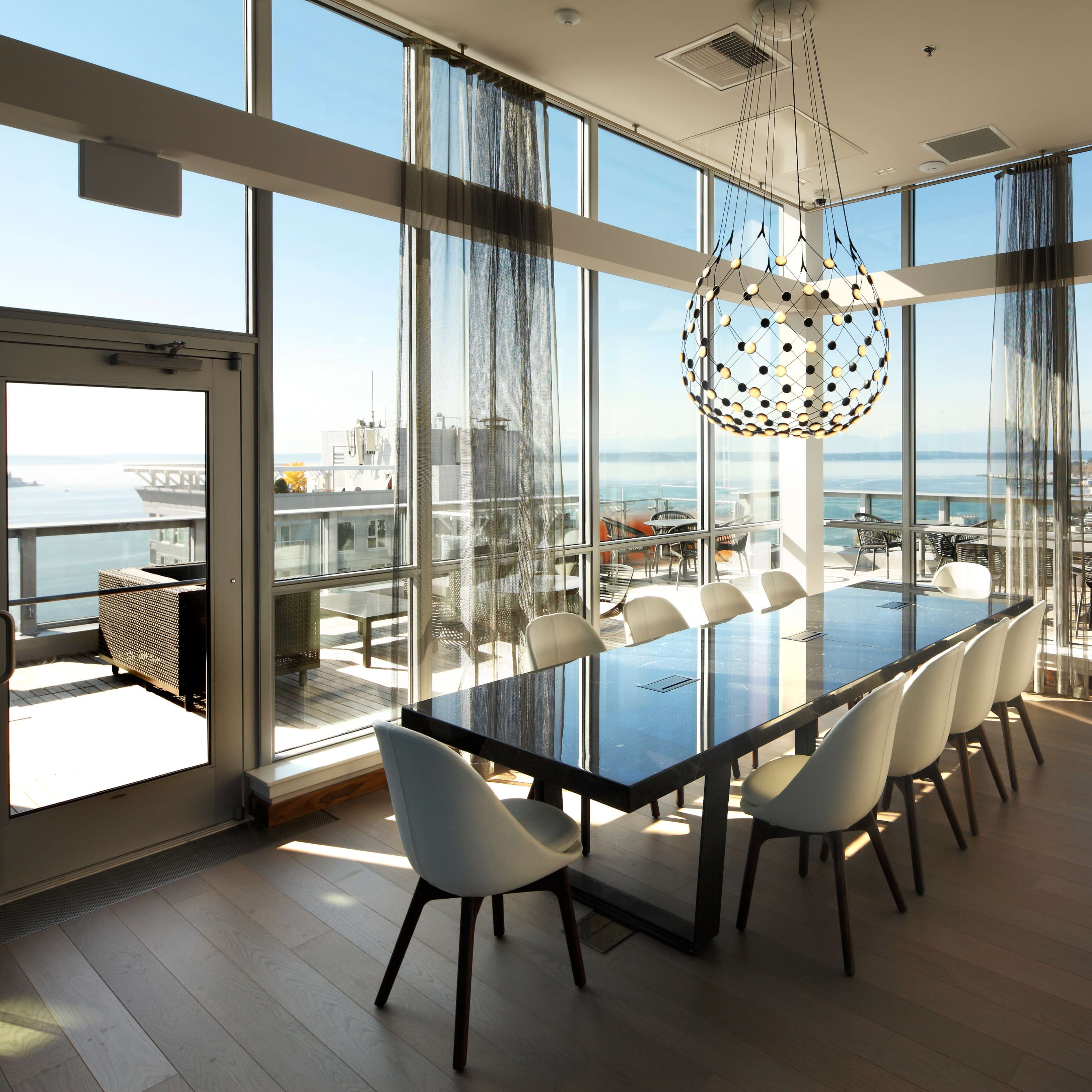A modern dining area features a long table surrounded by white chairs, with large windows offering a view of the water and city skyline.