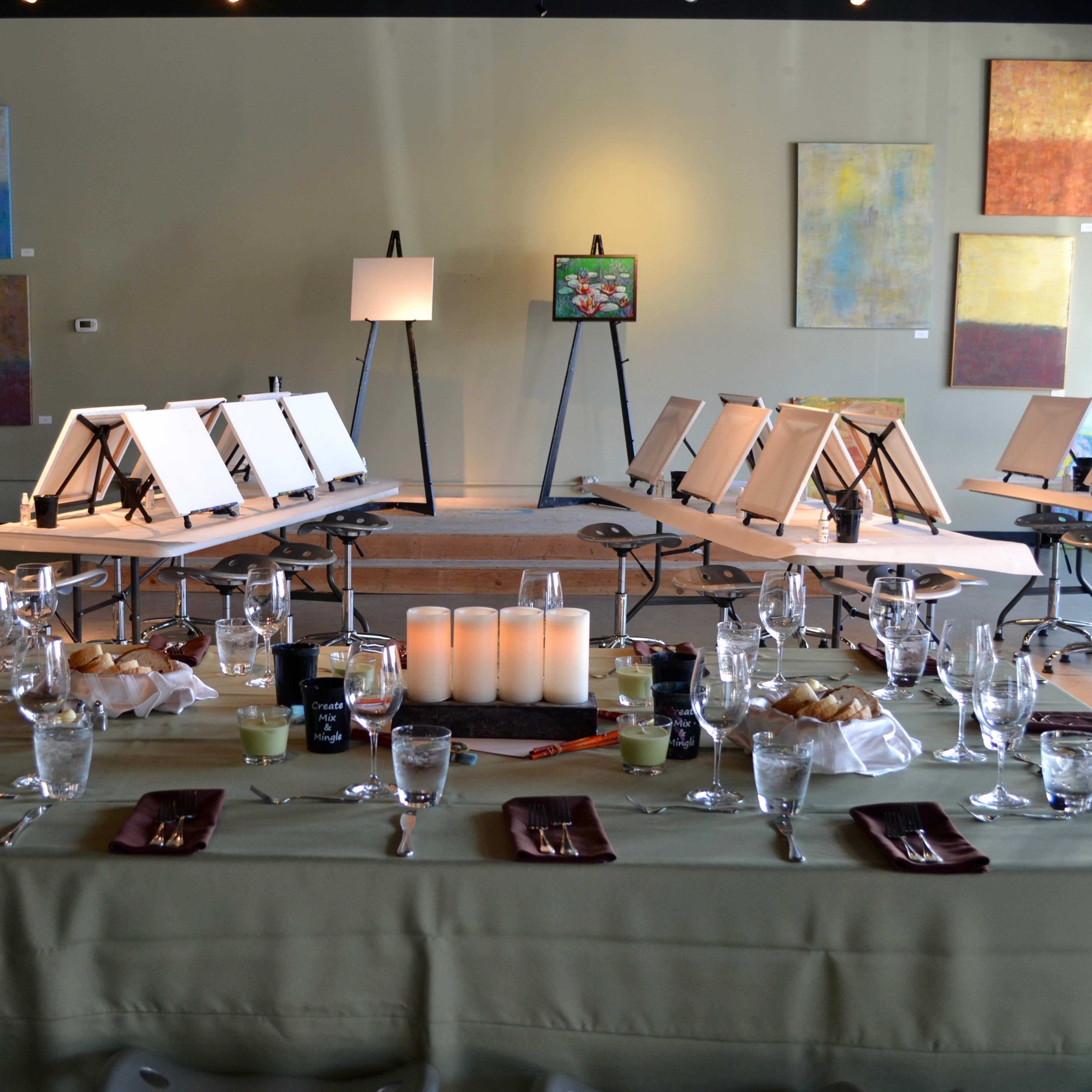 A spacious art studio features a long dining table set with glasses and plates, surrounded by easels with blank canvases.