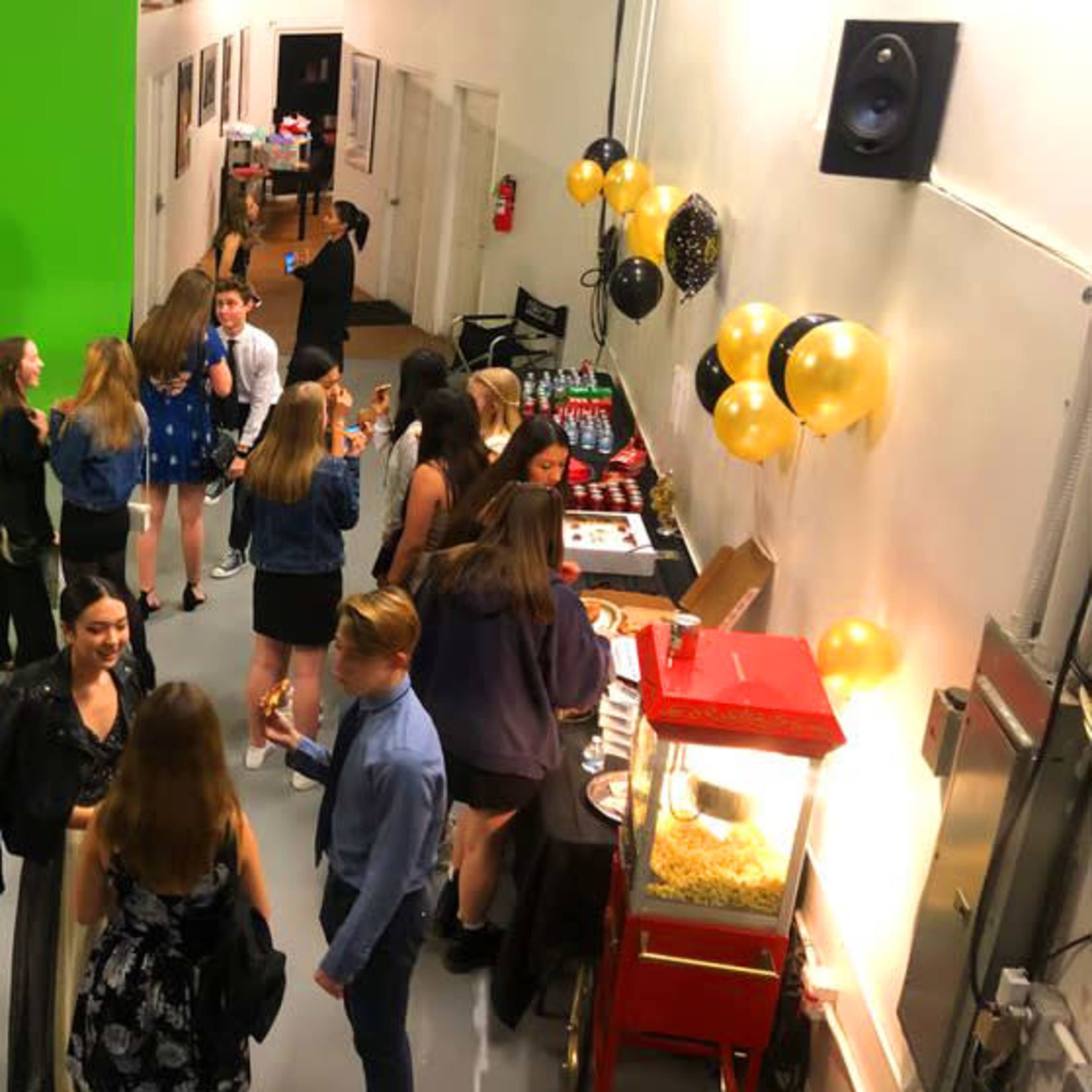 A group of people socialize at an event featuring a food table, a popcorn machine, and black and gold decorations.