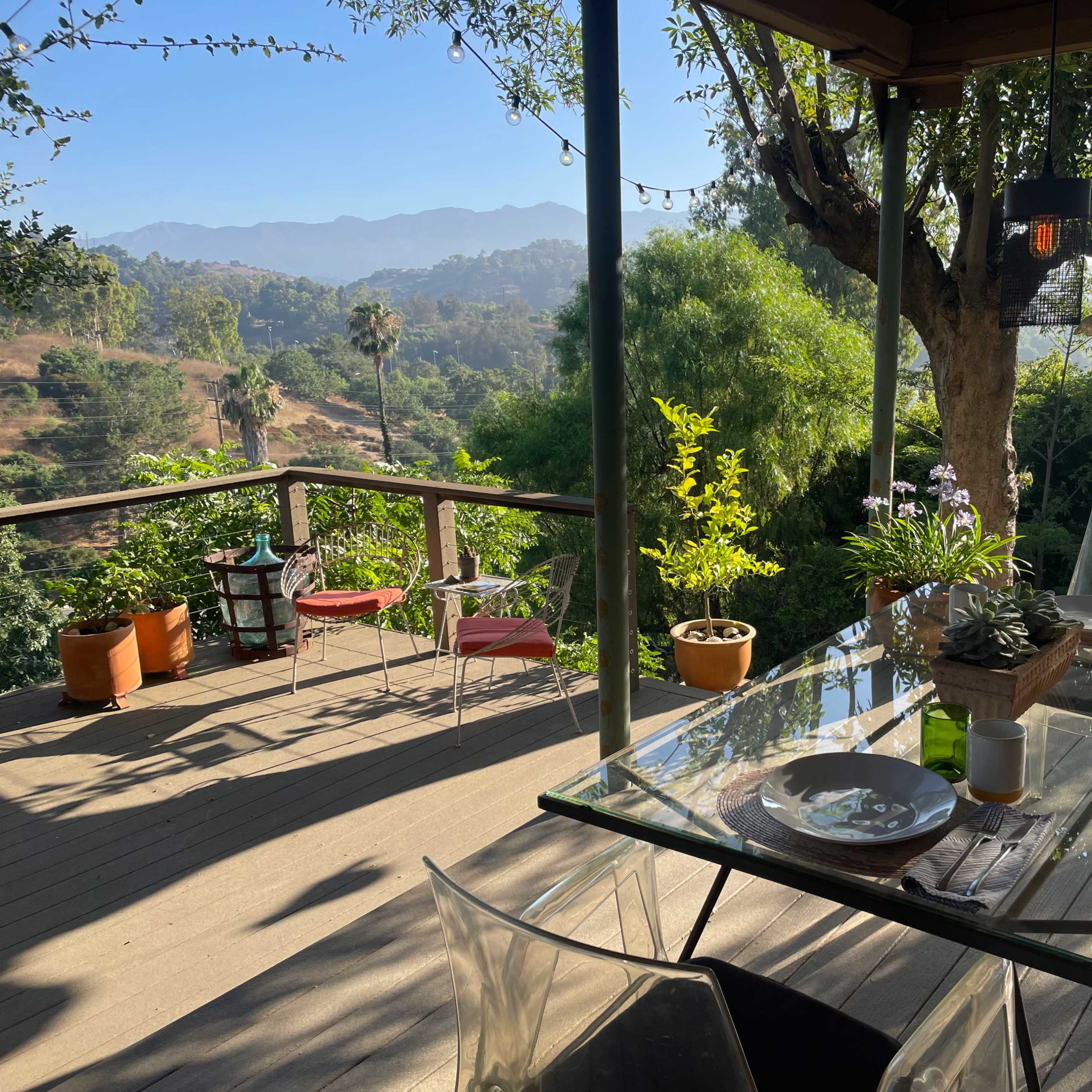 A glass dining table is set with plates on a deck overlooking a lush landscape with distant mountains.