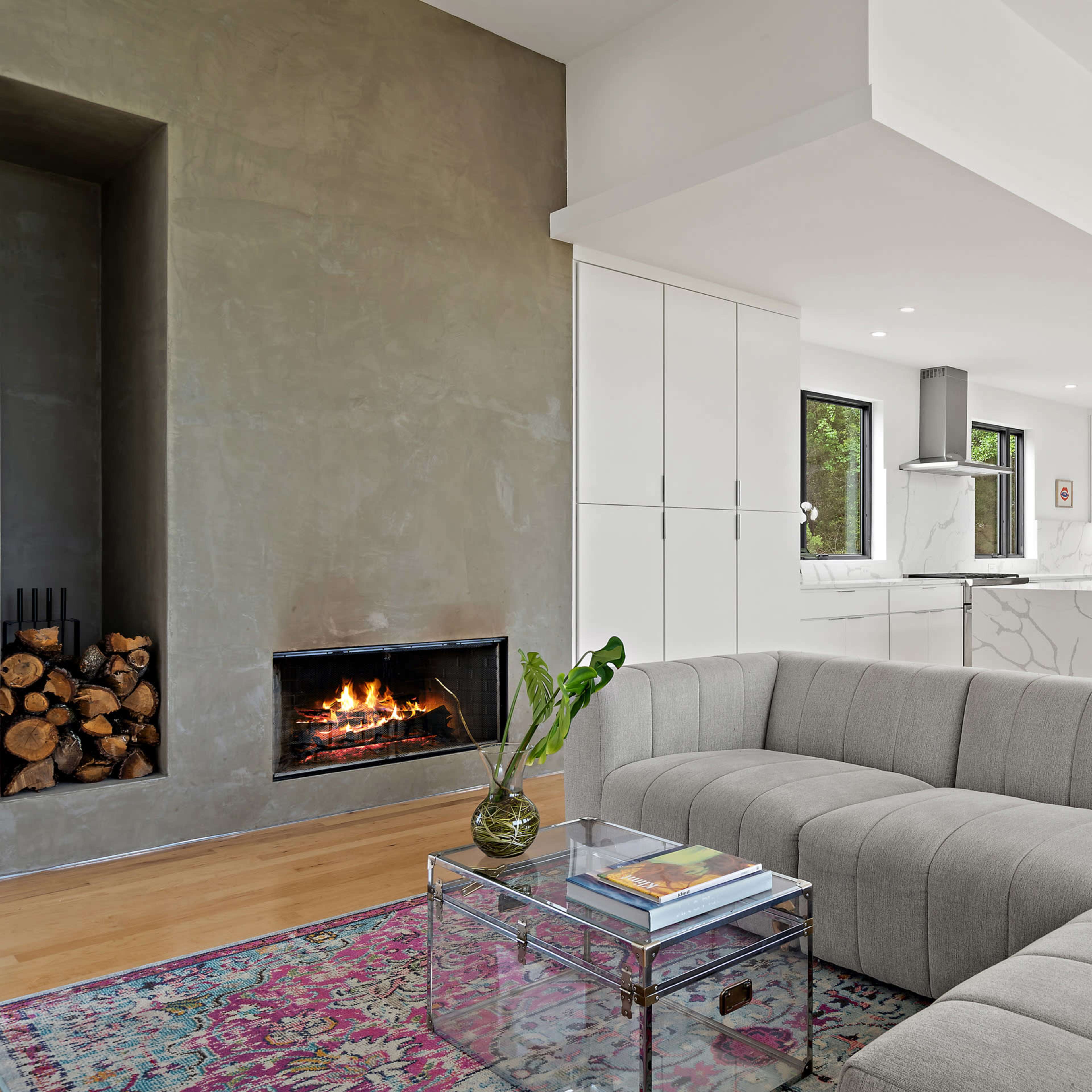 A modern living space features a gray sectional sofa, a glass coffee table, a wood-burning fireplace, and a view of an open kitchen.