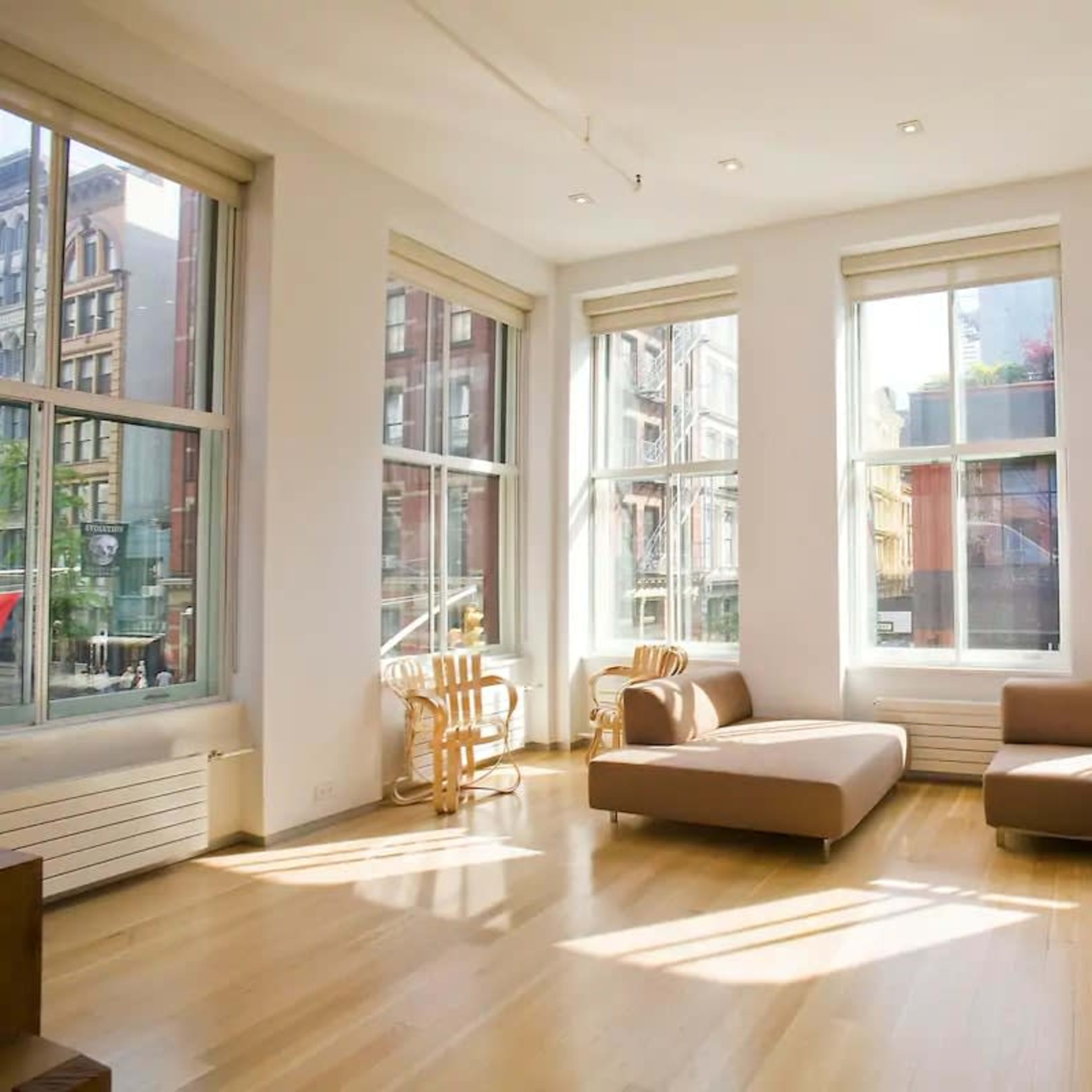 A bright living room features large windows, parquet flooring, and minimalist furniture including a sectional sofa and wooden chairs.