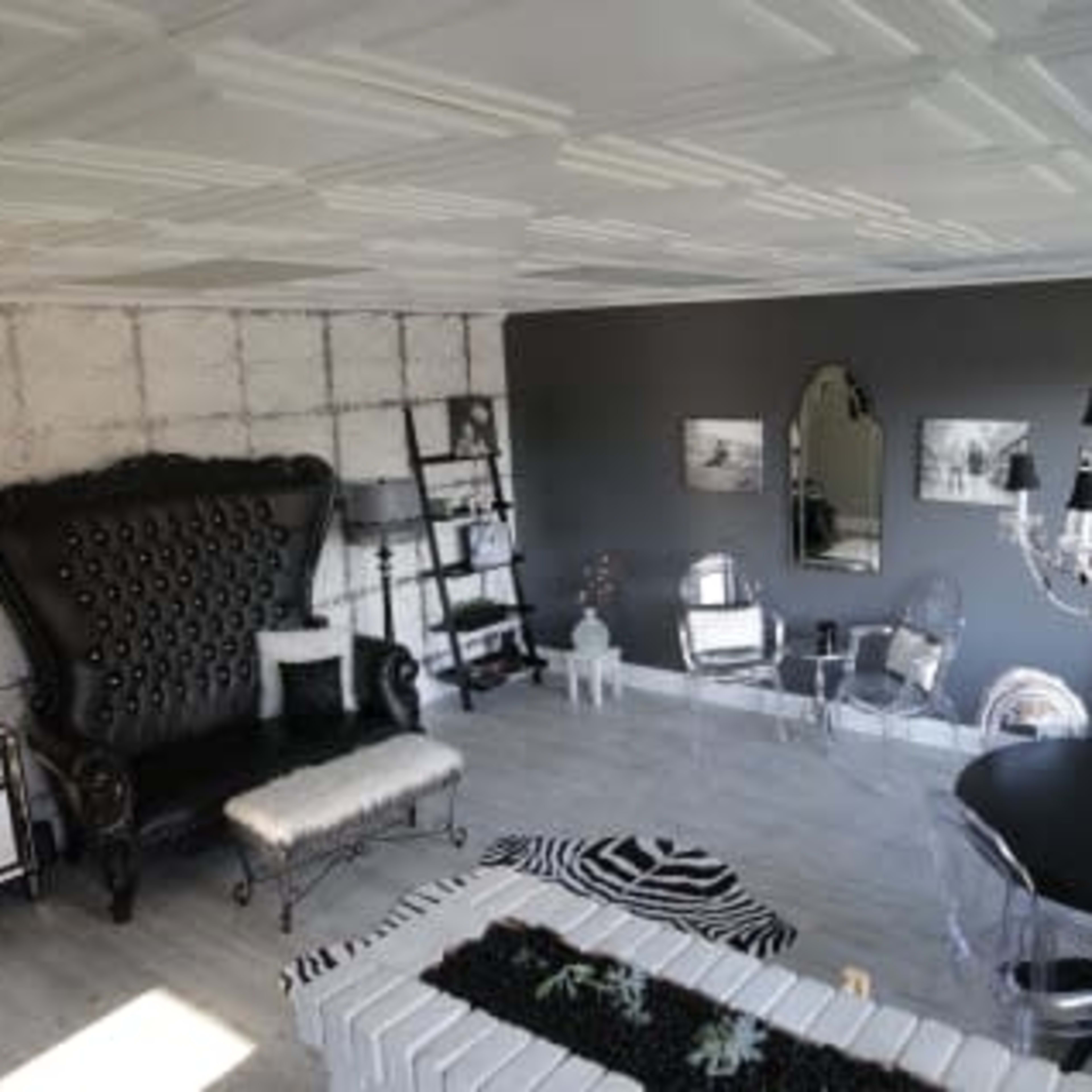 A modern living room featuring a large black tufted armchair, a mirrored shelf, clear acrylic chairs, a round black dining table, and a decorative chandelier against a textured wall.