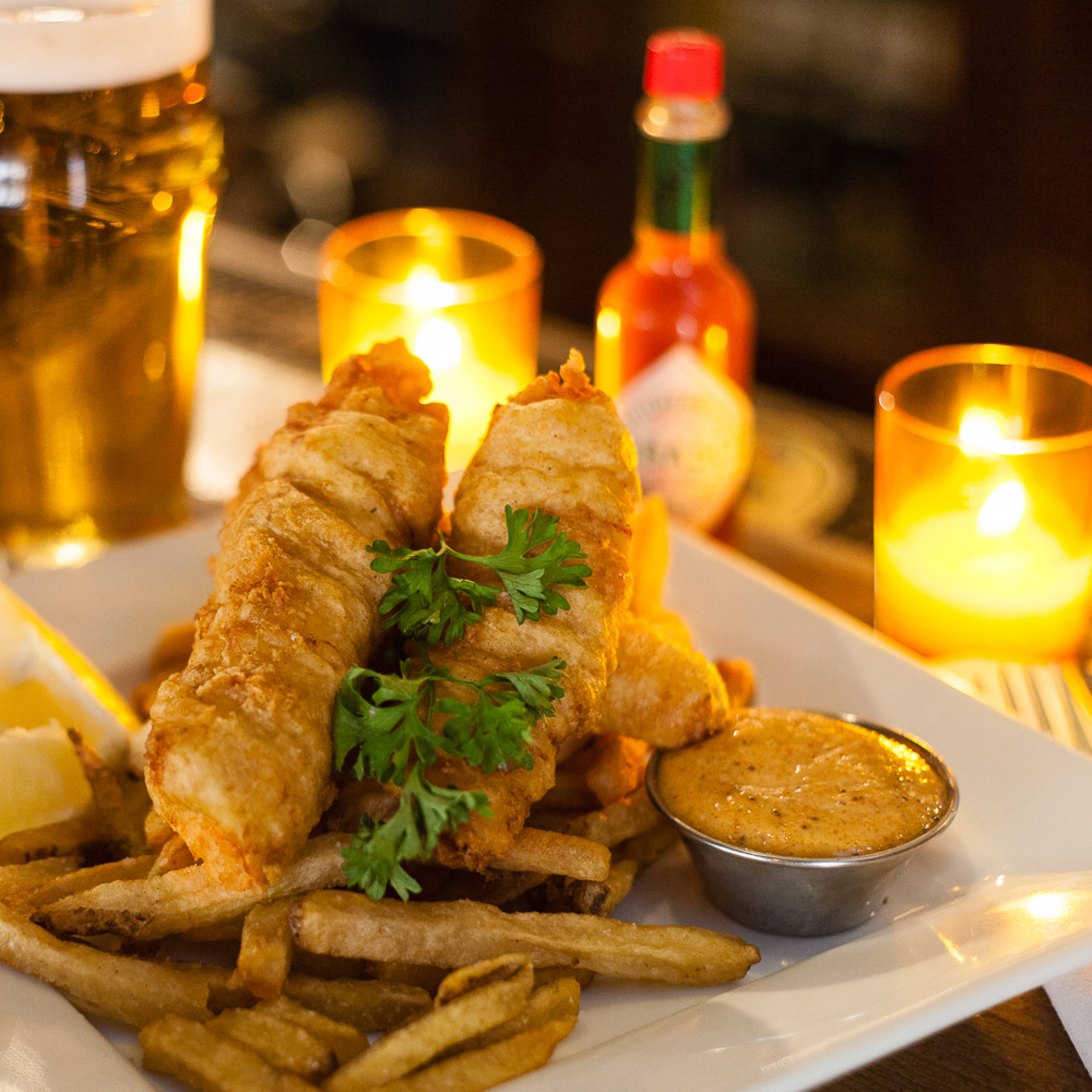 A plate of fish and chips is served with a side of dipping sauce, garnished with parsley, alongside a beer and condiments on a bar table.