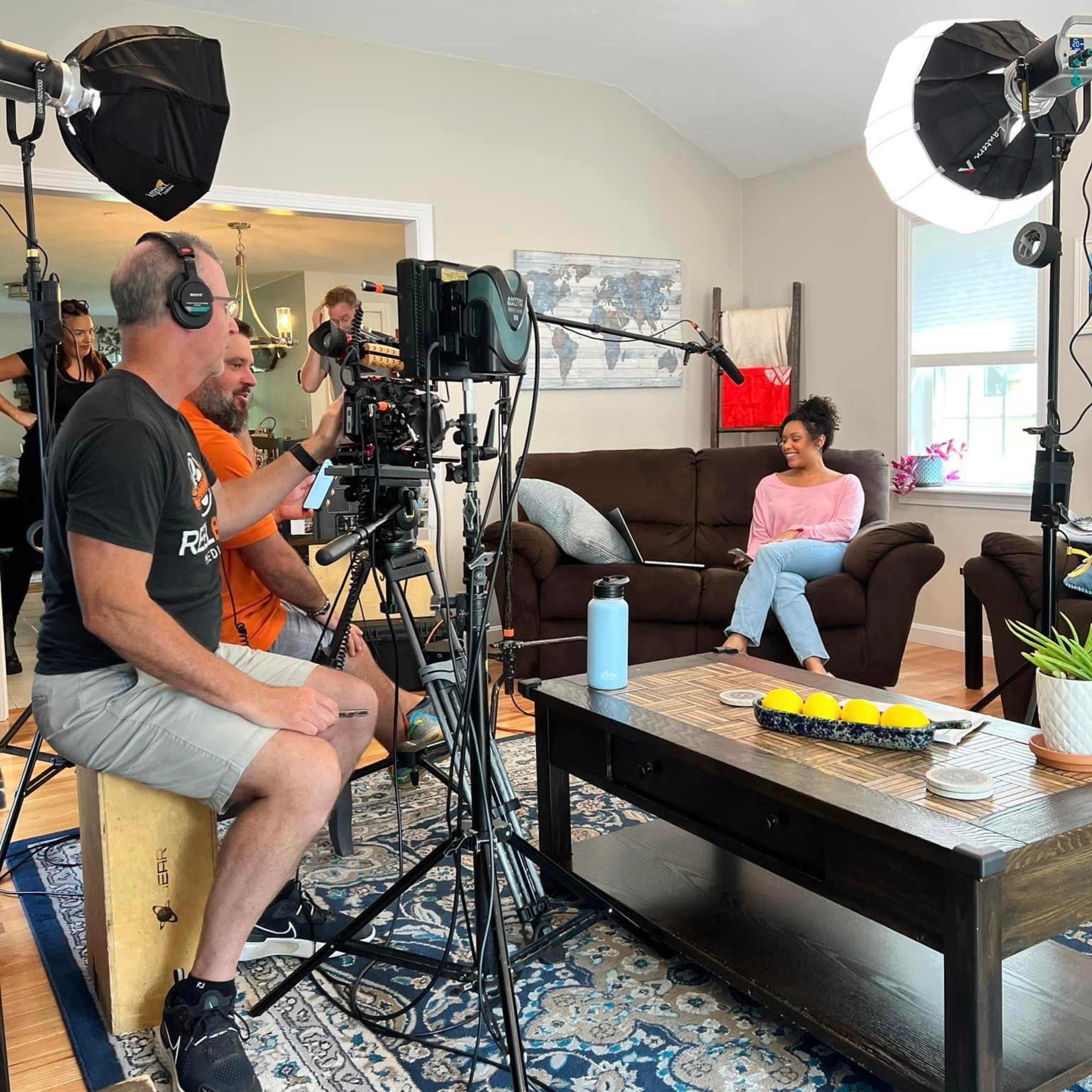 A film crew sets up cameras and lighting in a living room while a woman sits on a couch preparing for an interview.