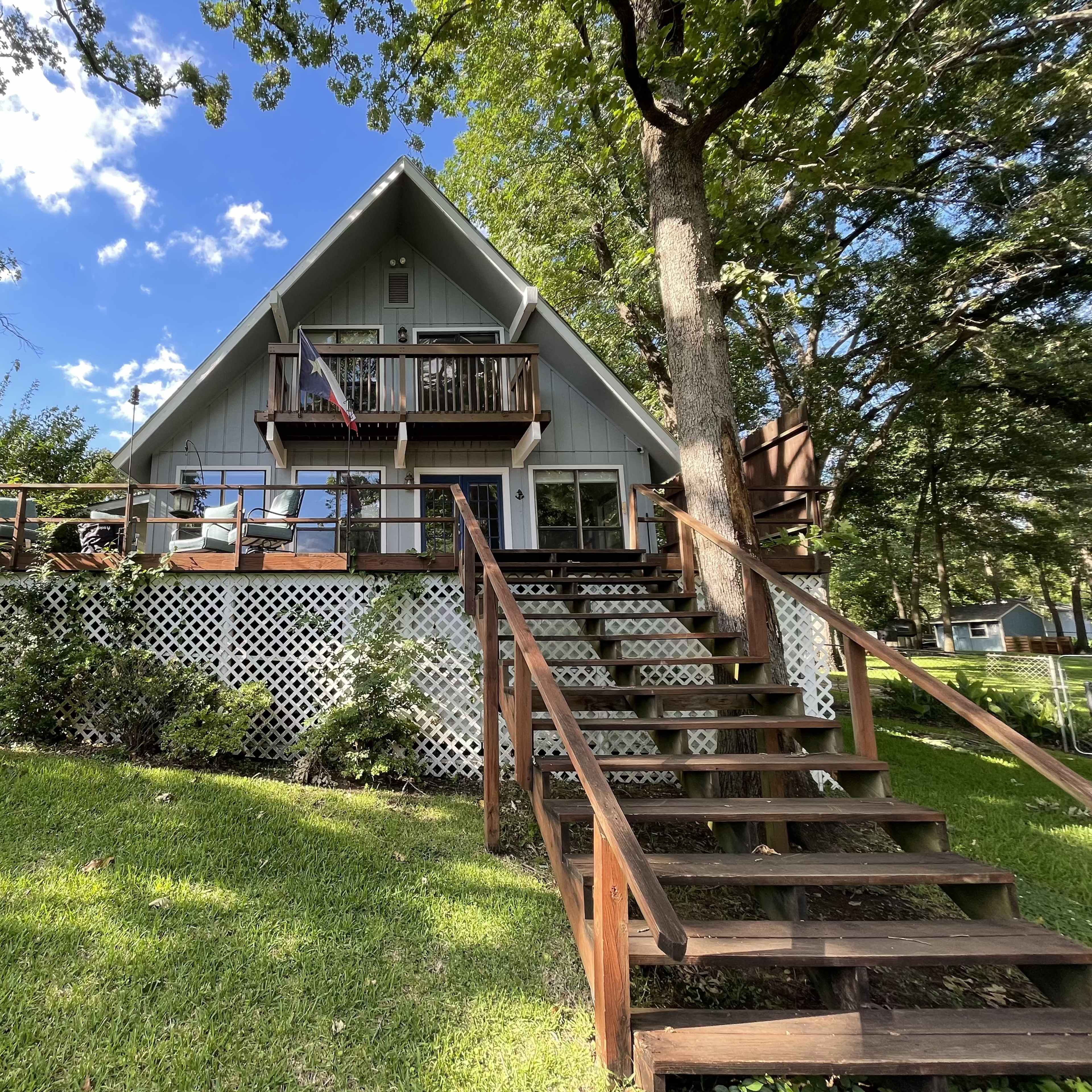 A two-story A-frame cabin with a wooden deck and stairs, surrounded by trees and lawn, is set against a blue sky with scattered clouds.