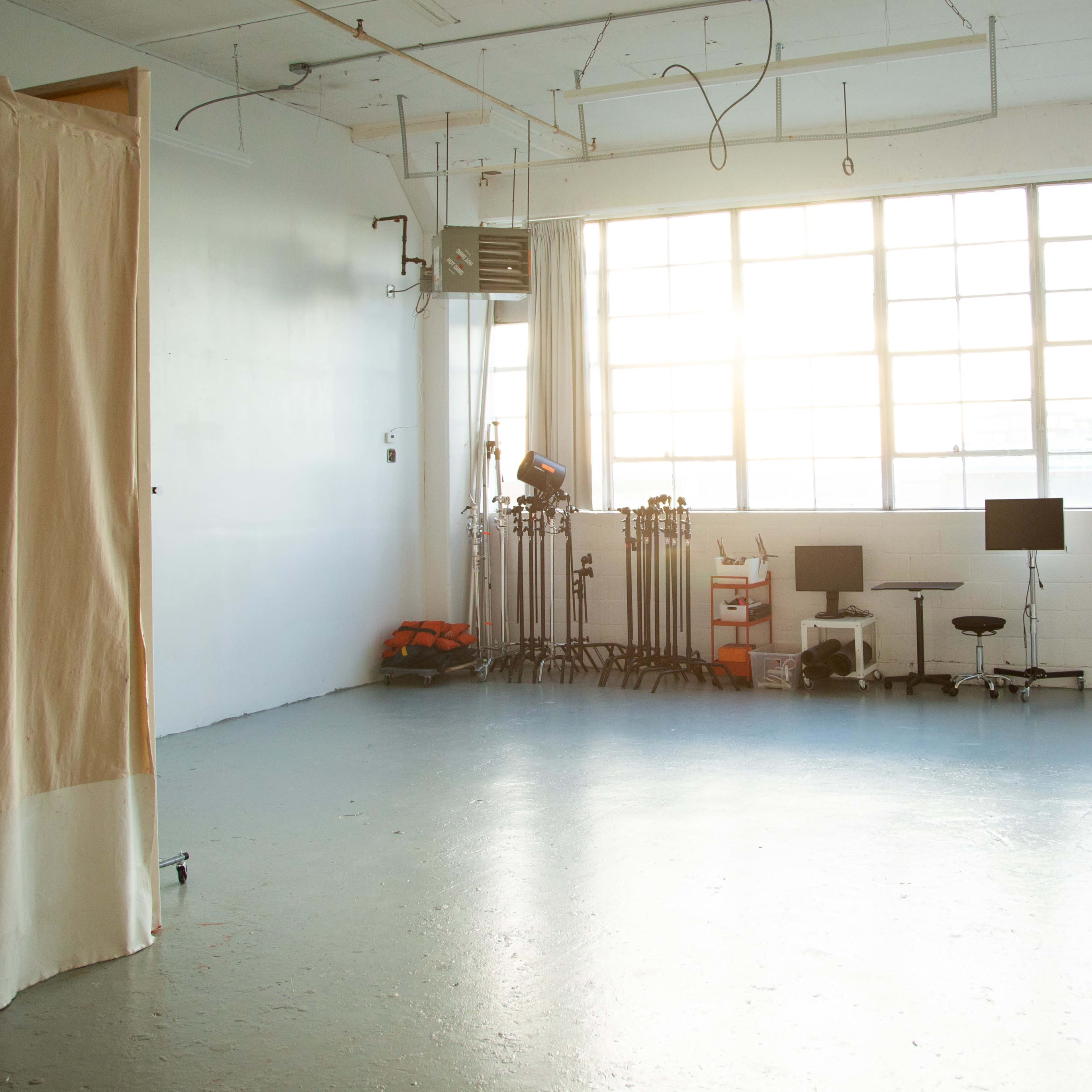An empty studio space with large windows, beige curtains on one side, and equipment set up including two computer monitors and stands.
