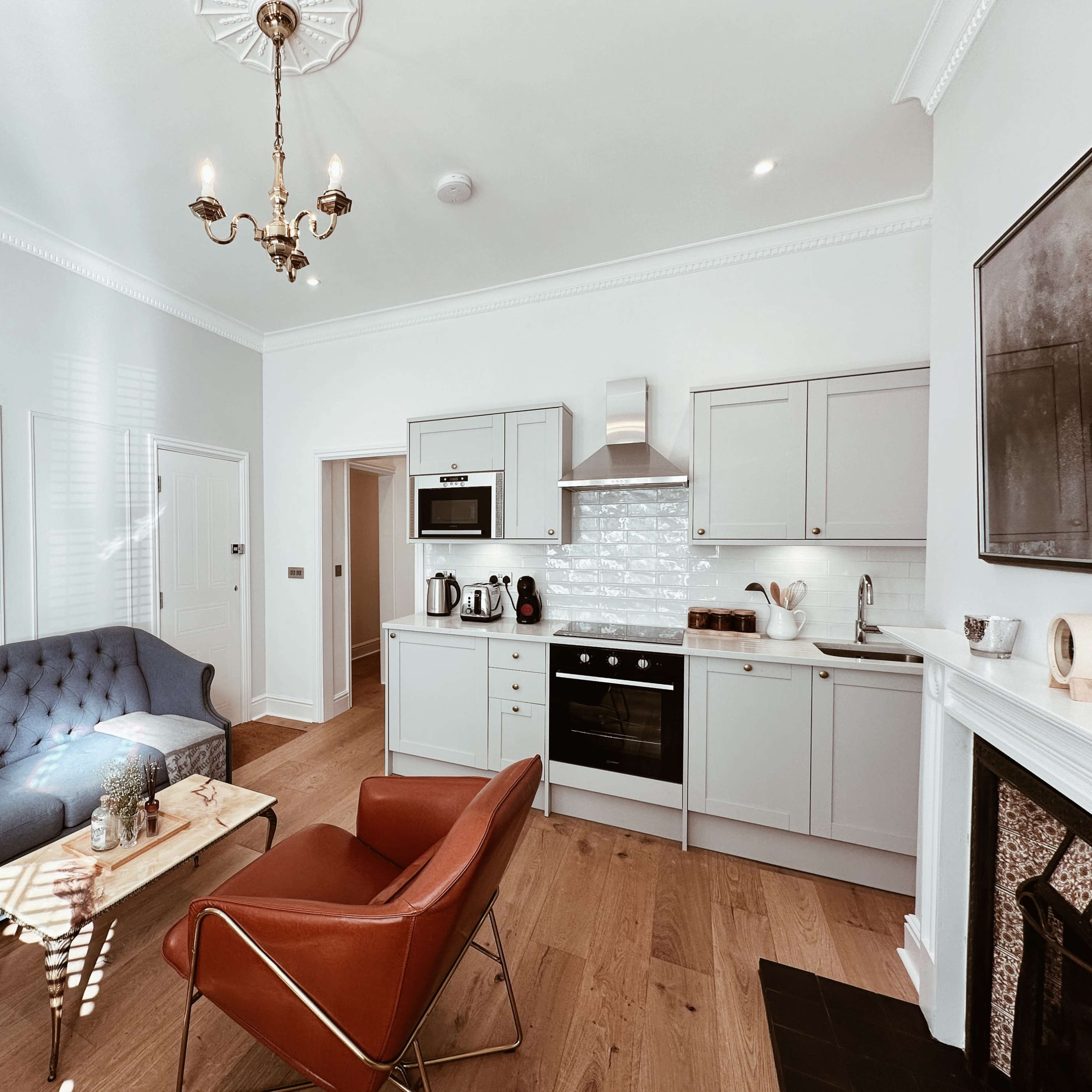 A modern living room with a blue tufted sofa, a red armchair, a wooden coffee table, and a kitchen area featuring light cabinetry and stainless steel appliances.