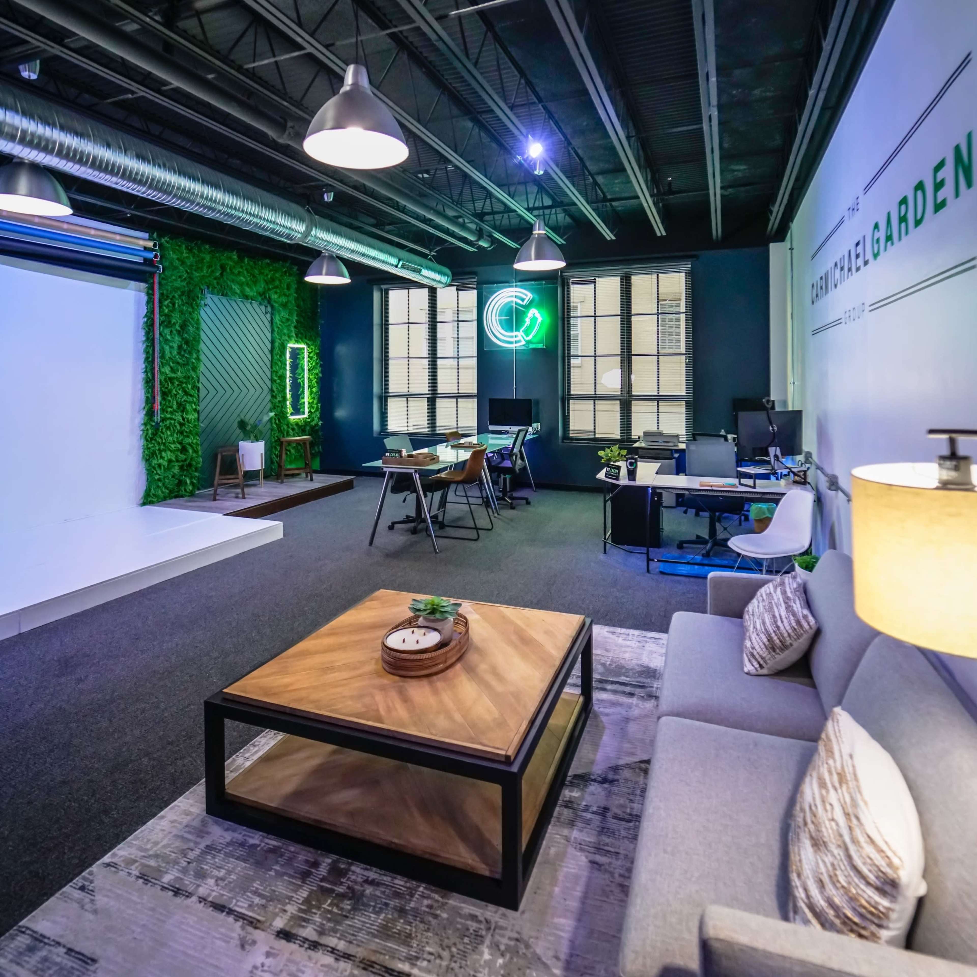 A modern office space featuring a lounge area with a gray sofa and a wooden coffee table, alongside workstations and greenery, illuminated by overhead lighting.