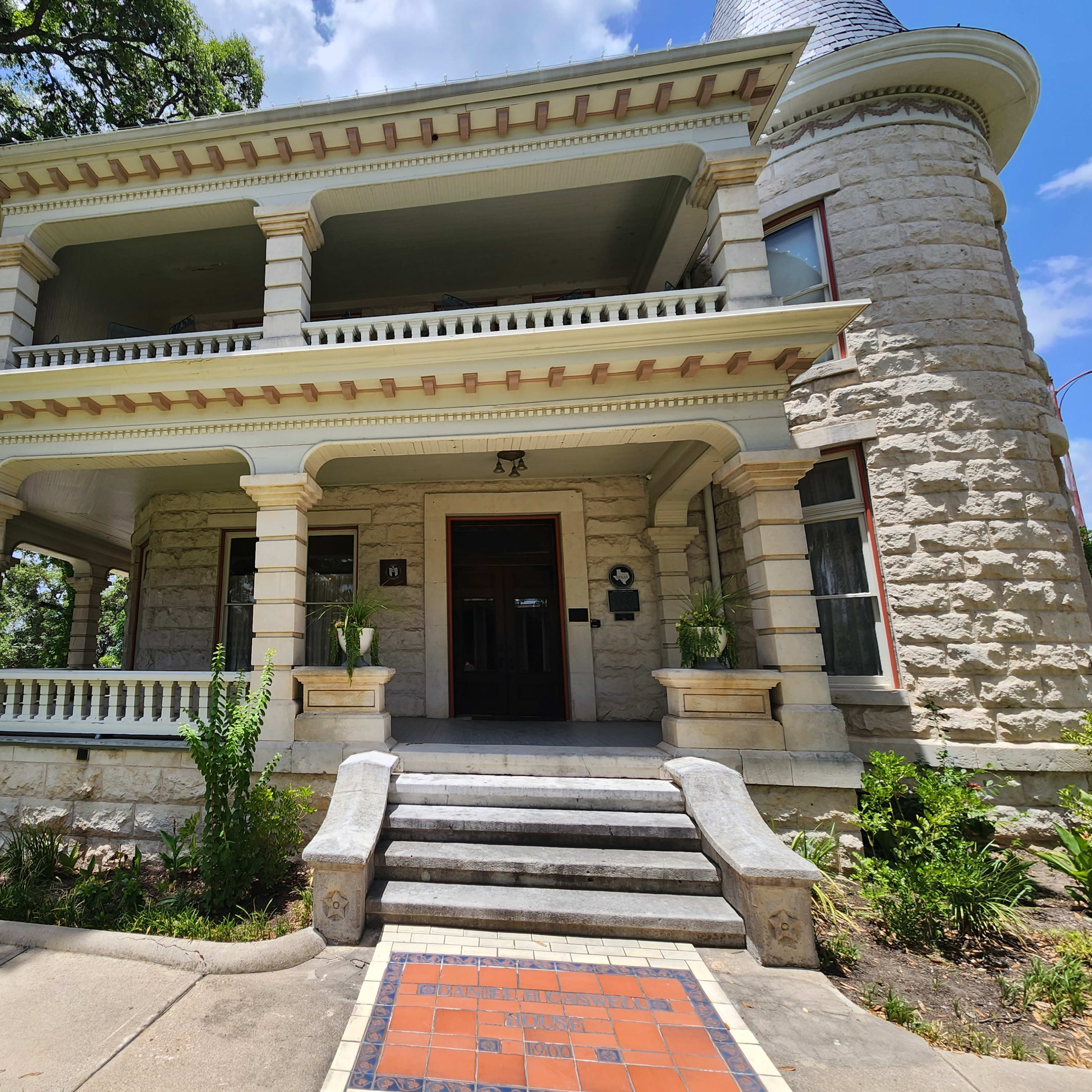 A historic stone building with two levels, featuring a grand entrance and decorative columns, surrounded by greenery and a pathway leading to the front steps.