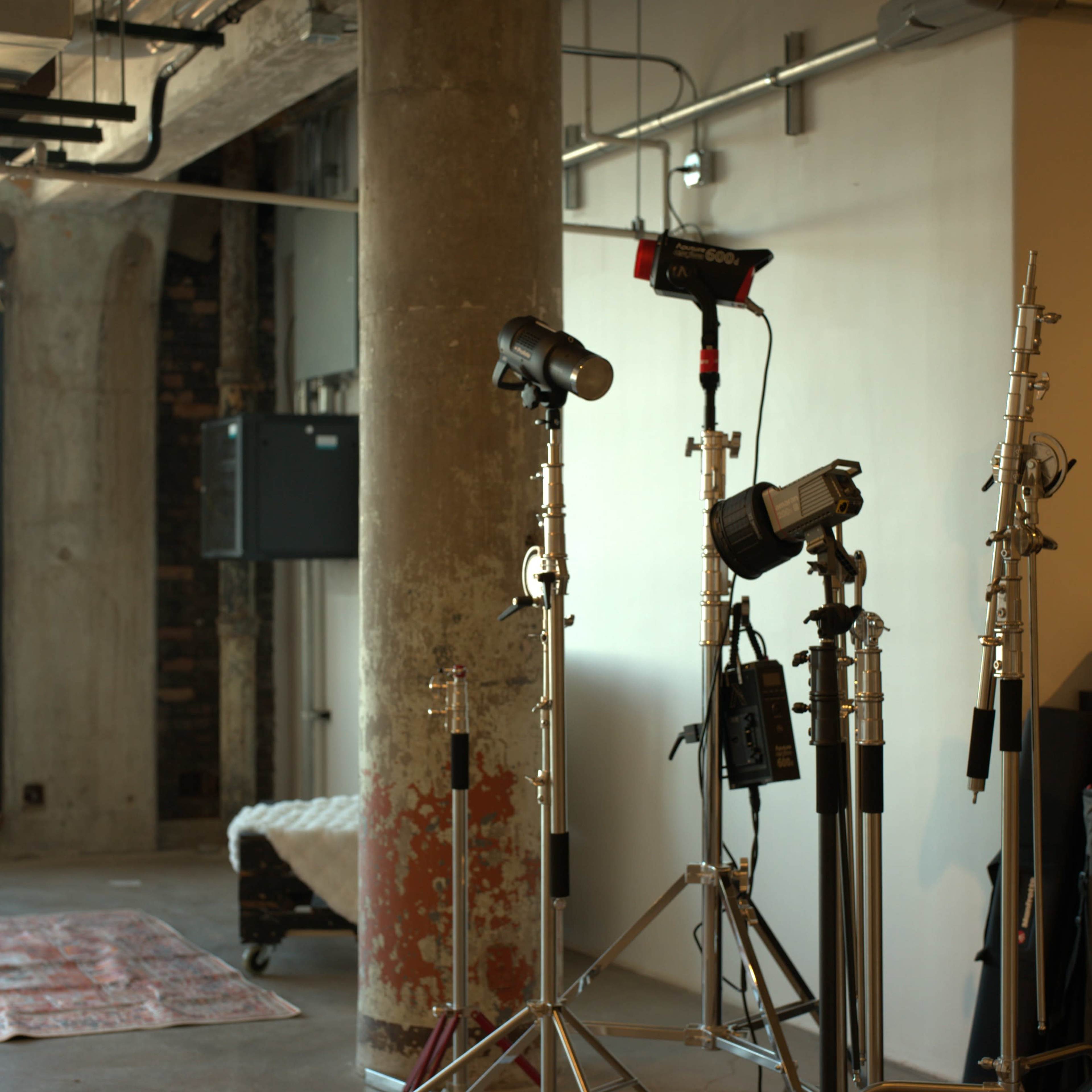 A studio space with lighting equipment set up near a concrete pillar and a partially visible area with a patterned rug.