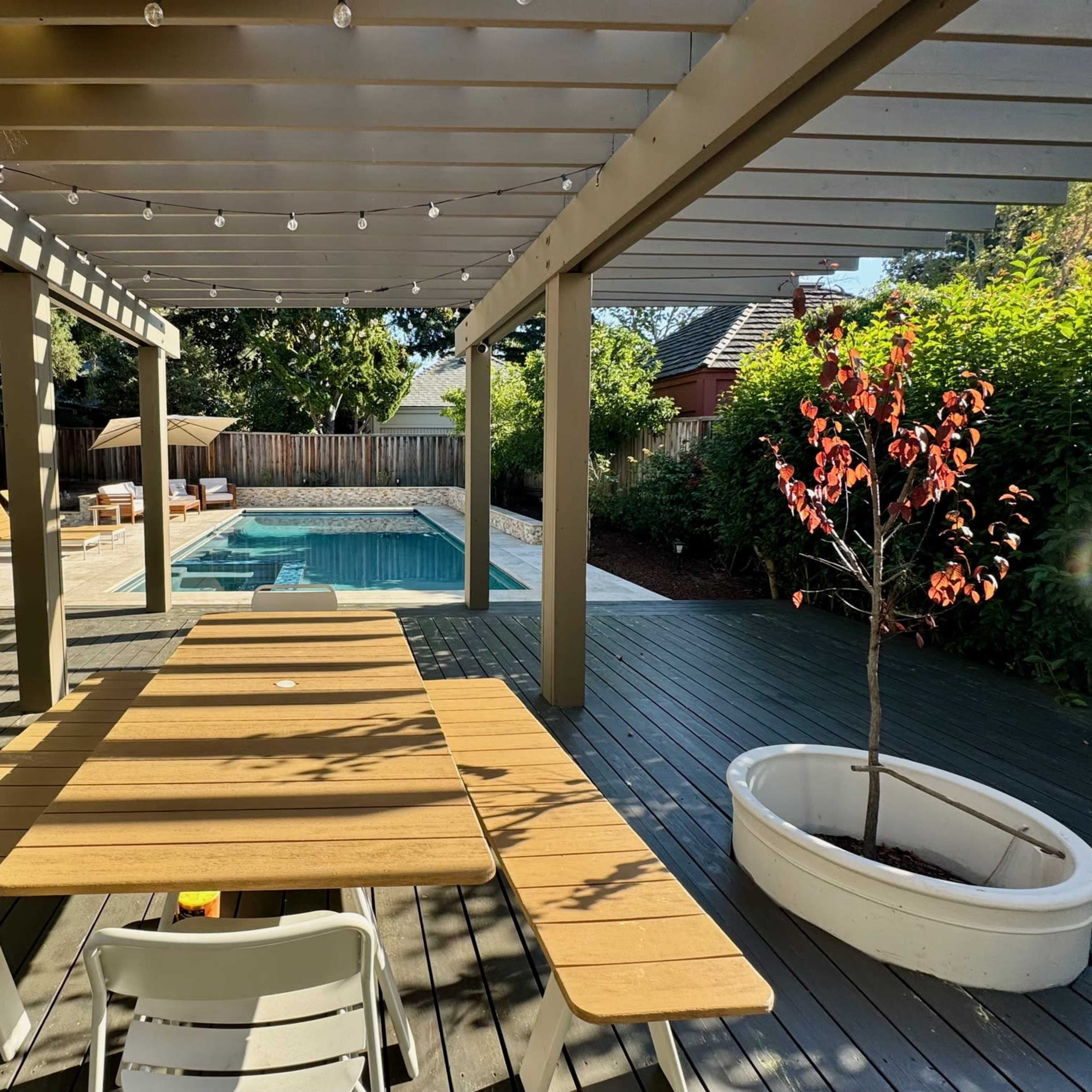 A wooden table sits under a pergola, overlooking a swimming pool surrounded by greenery and a wooden fence.