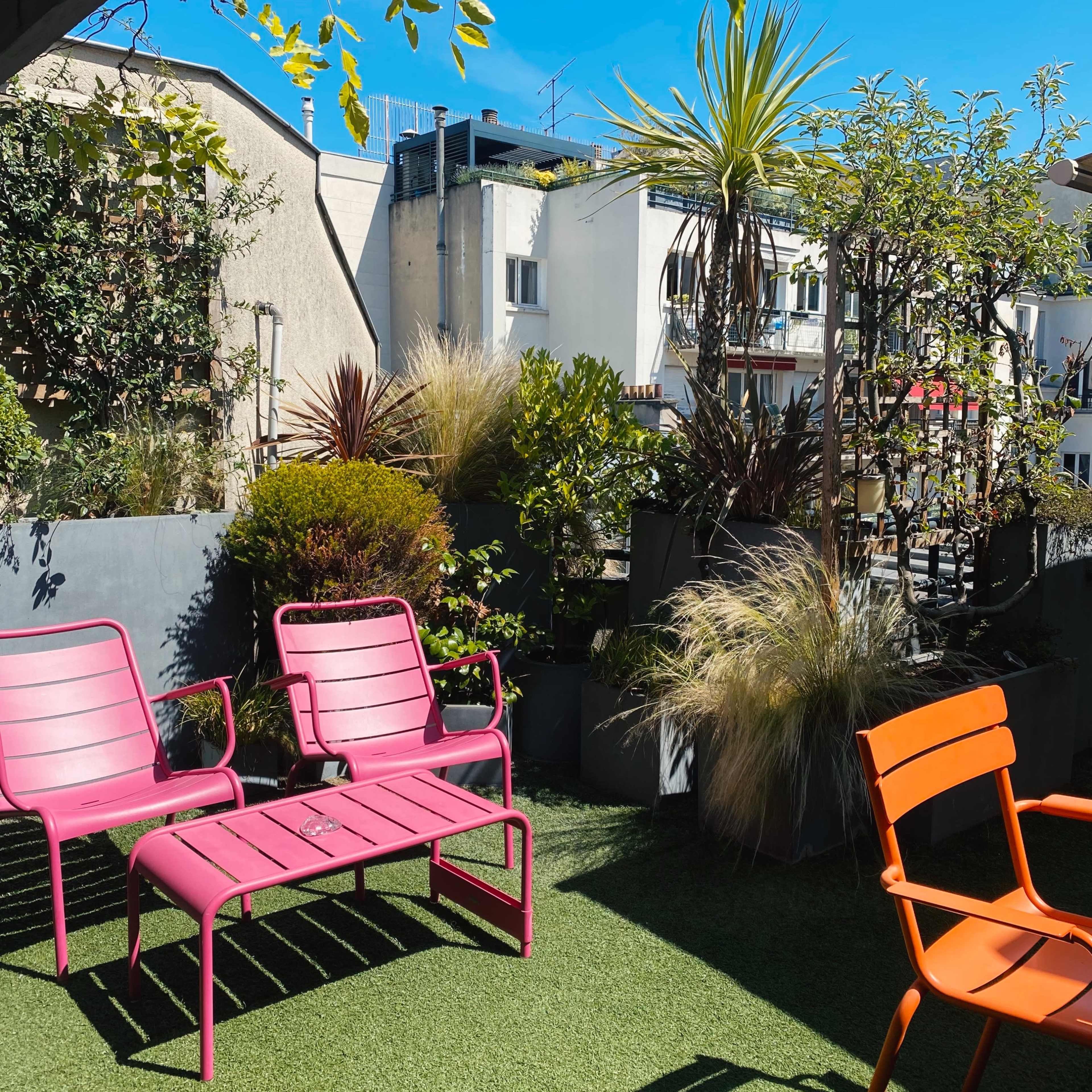 A rooftop garden featuring pink and orange lounge chairs, surrounded by various plants and greenery against a backdrop of buildings and a clear blue sky.