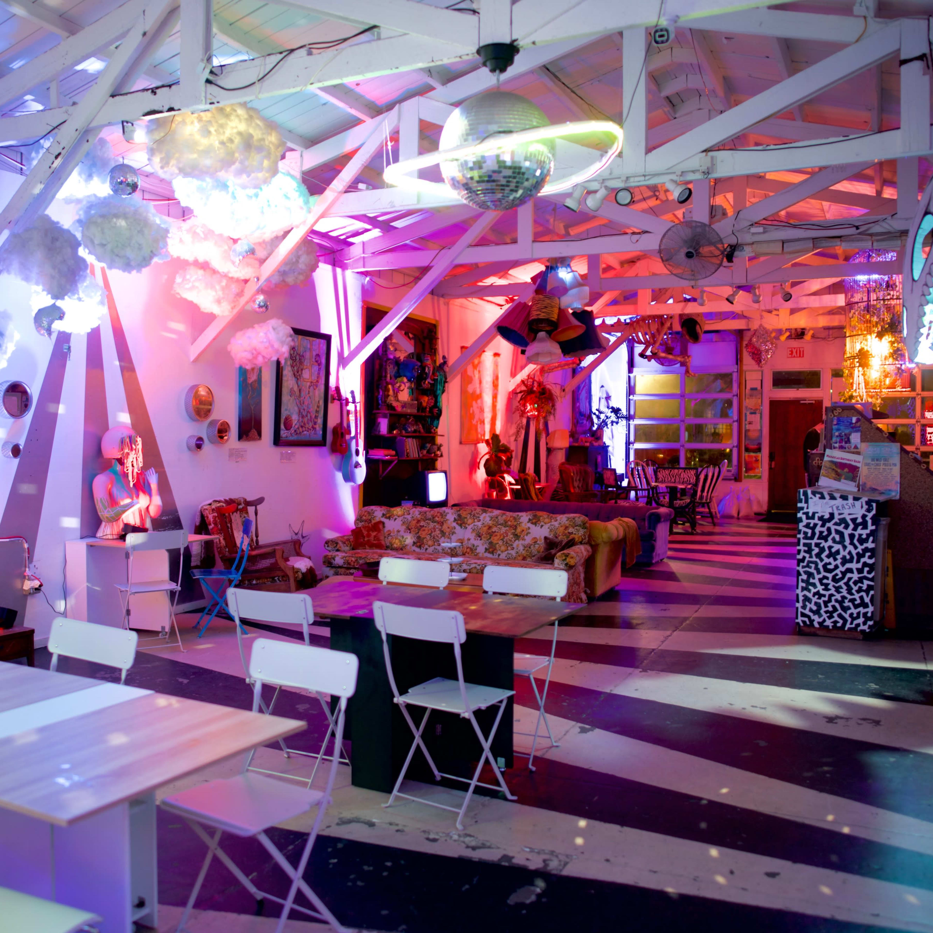 A vibrant room with colorful decor featuring white tables and chairs, artwork on the walls, and whimsical cloud-like installations hanging from the ceiling.