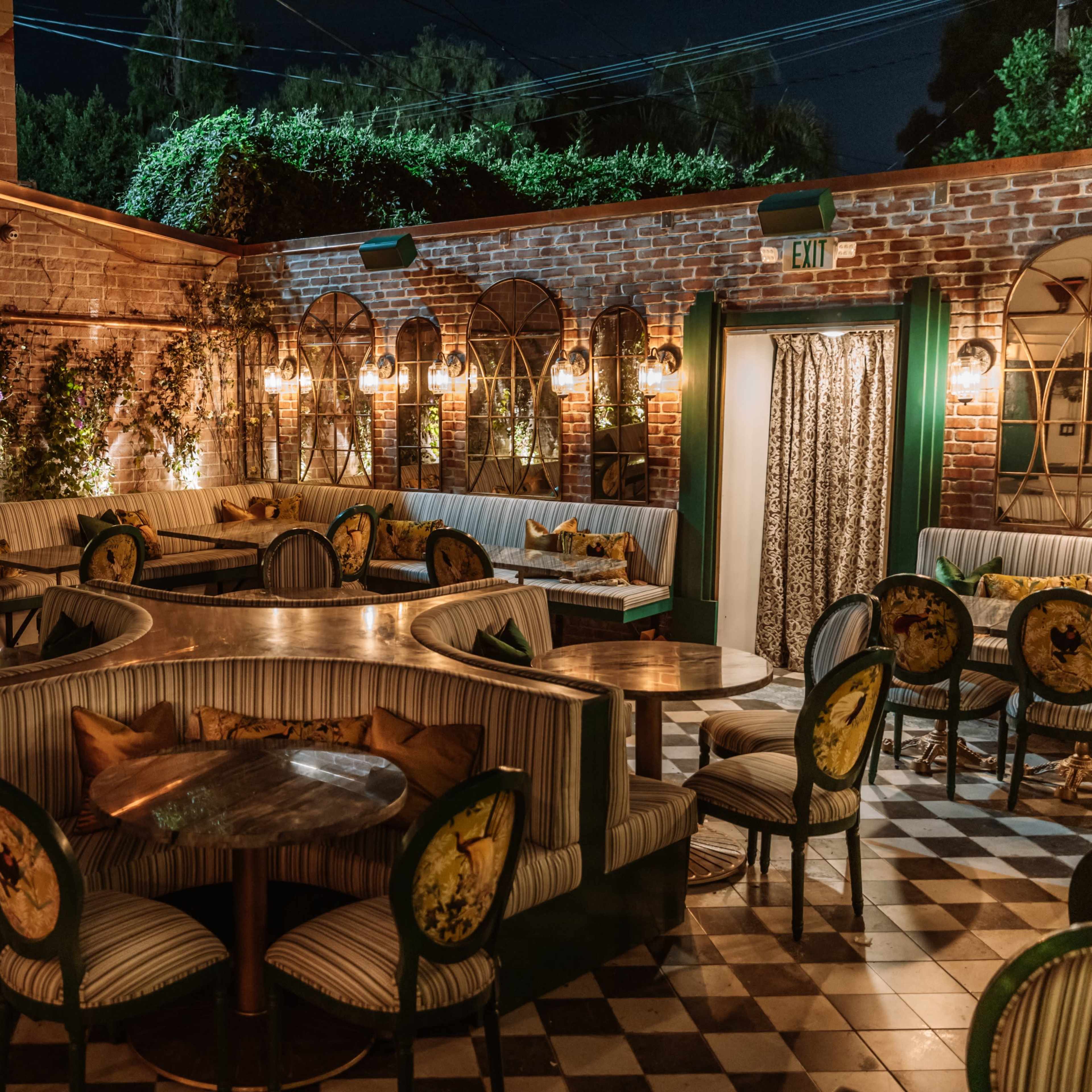 A dimly lit, elegant restaurant patio with striped seating, wooden tables, and patterned decor on the walls, surrounded by greenery.