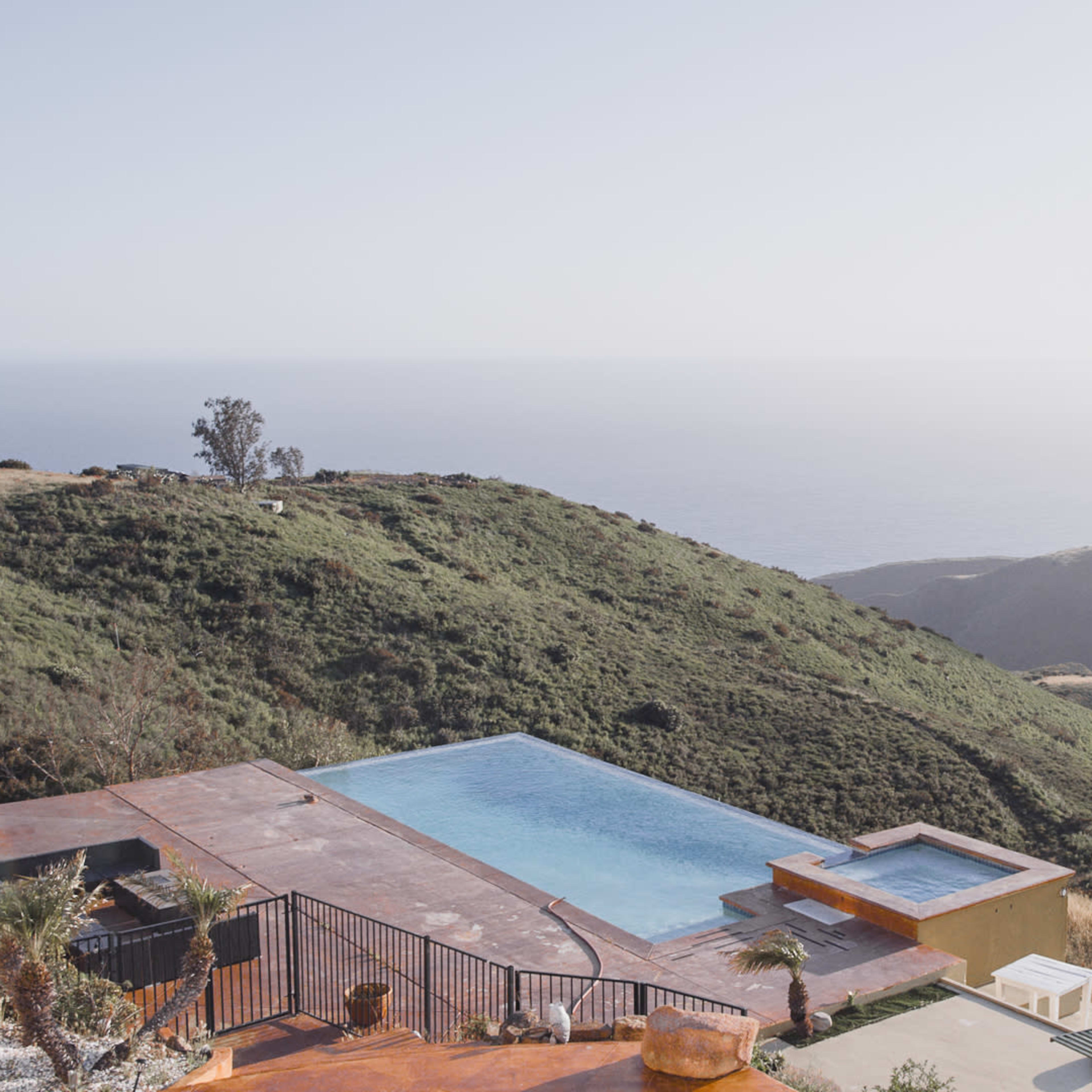 A modern infinity pool overlooks a hillside that slopes down to the ocean.