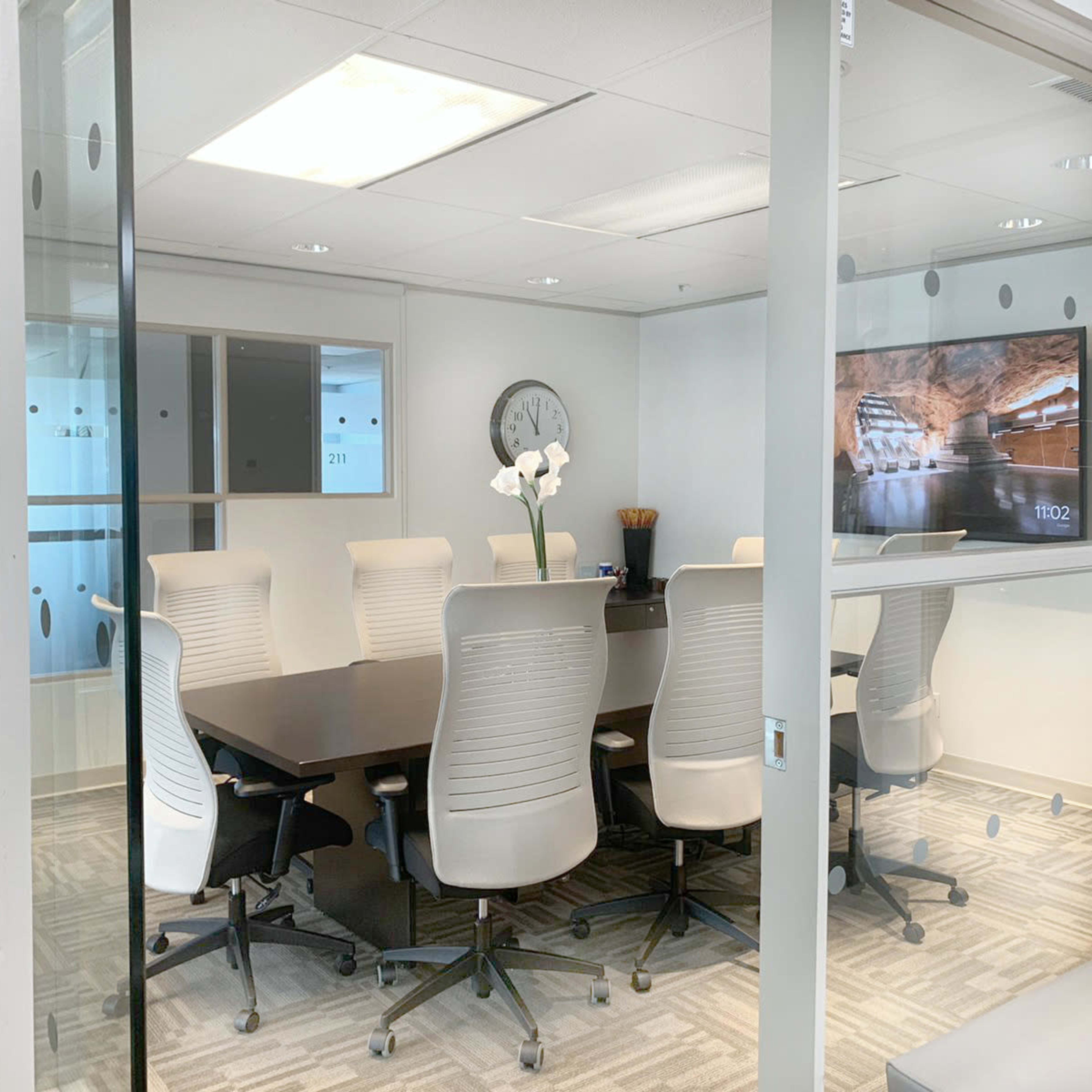 A glass-walled conference room features a large table surrounded by ergonomic chairs and a television displaying a scenic image.