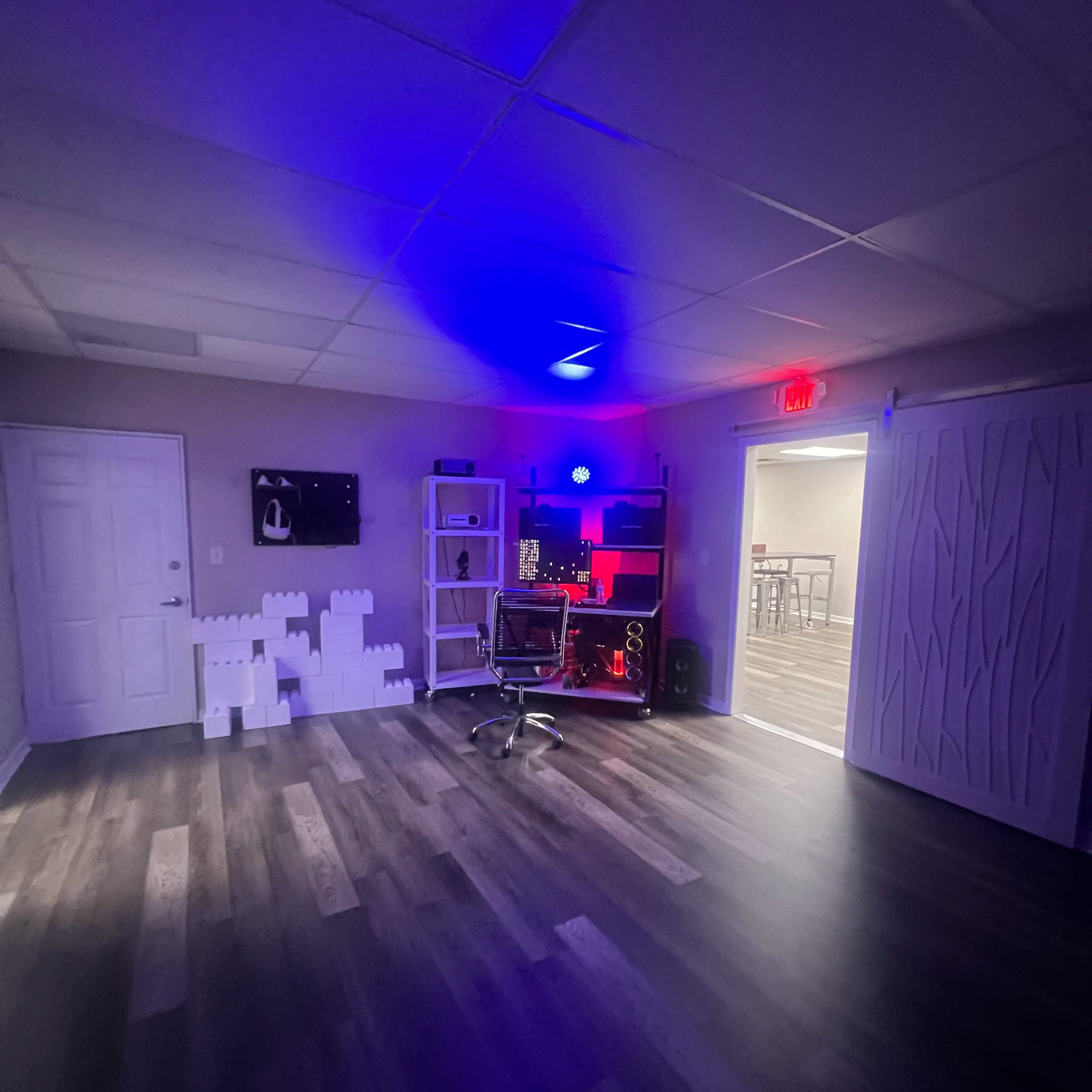 A spacious room features a desk with a computer setup, a chair, and decorative shelves, illuminated by blue and red lights, with a white textured wall and an open door.