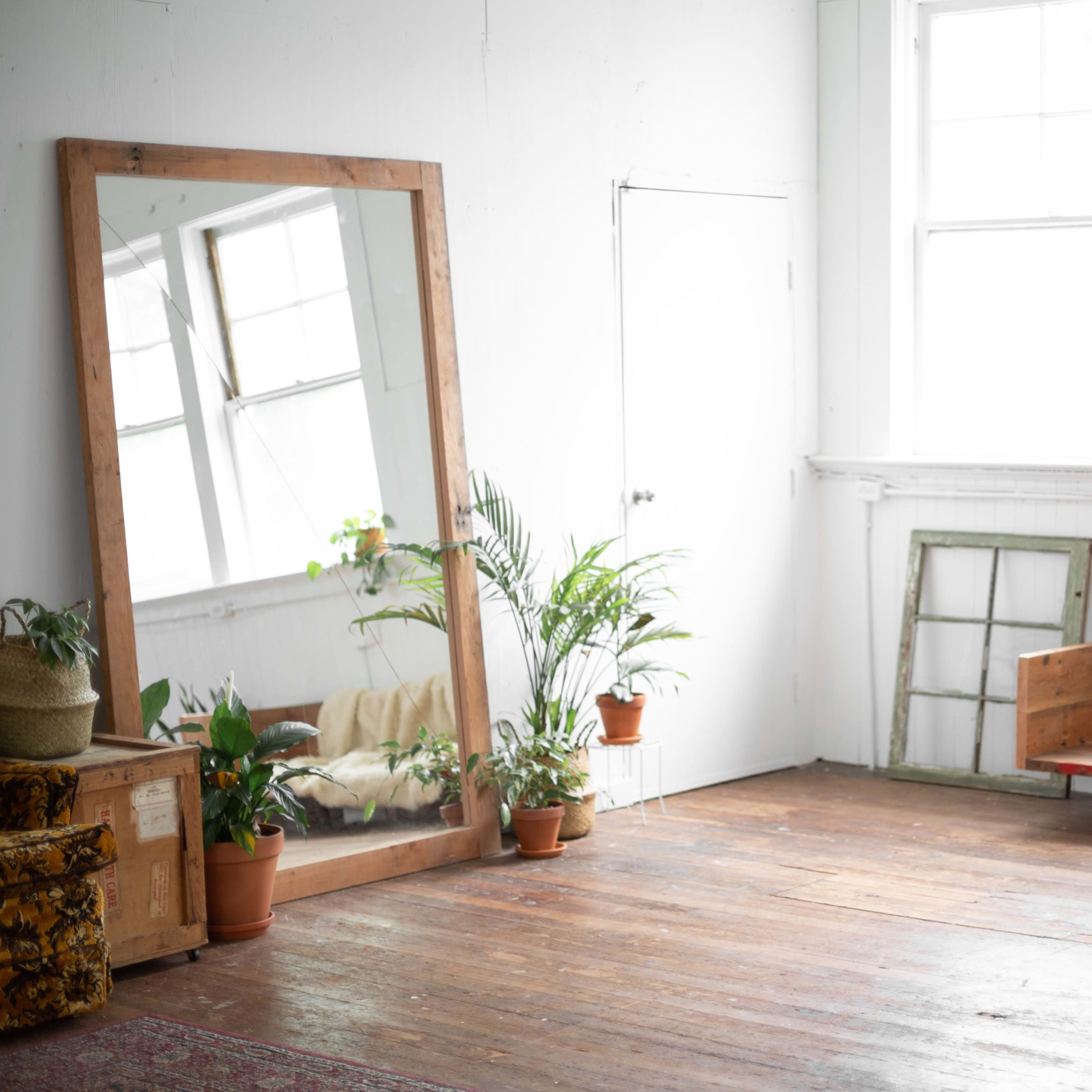 A spacious room features a large mirror, a wooden sofa with a blanket, a round orange ottoman, and several potted plants.