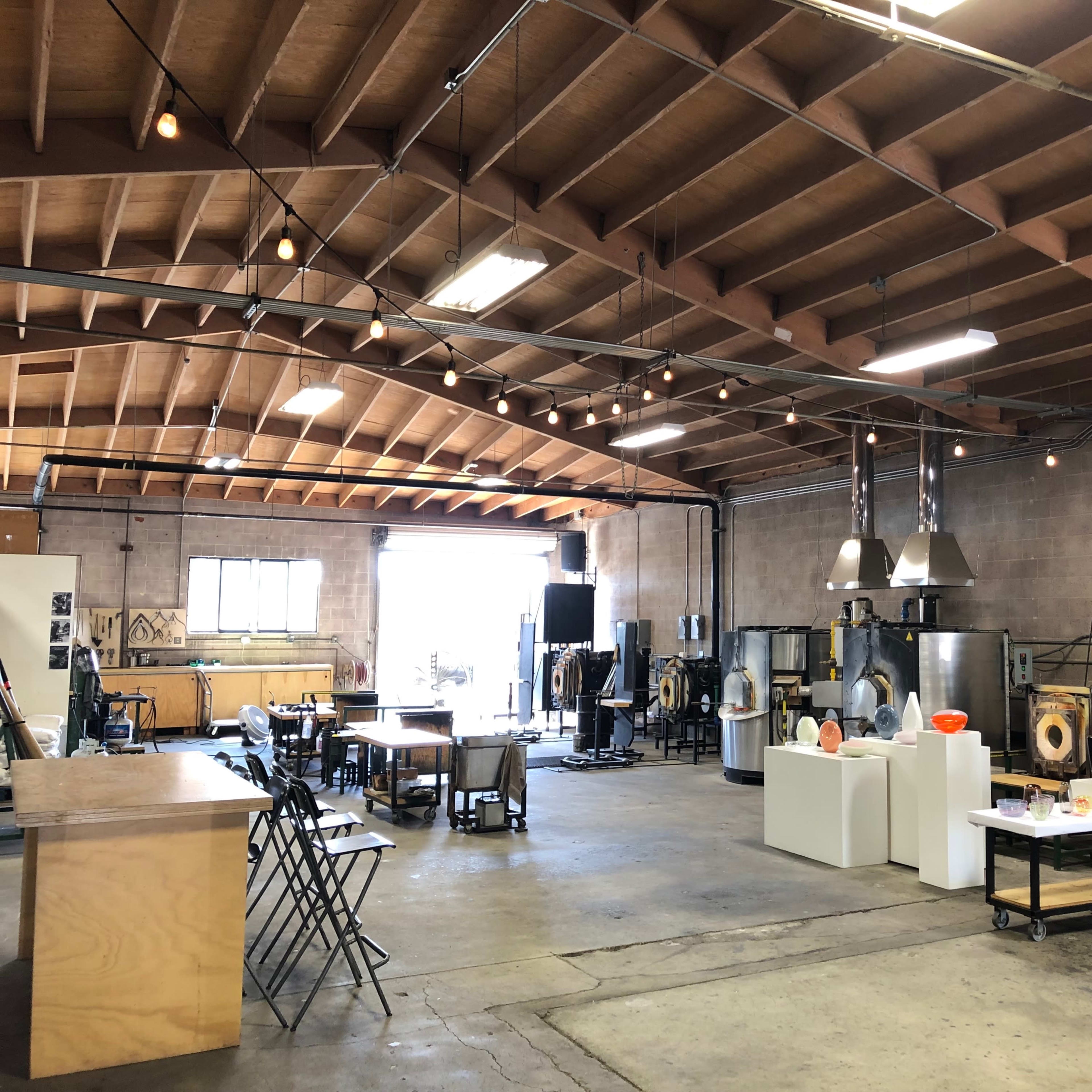 A spacious art studio with industrial equipment, tables, and display stands for glassblowing or similar craft activities.