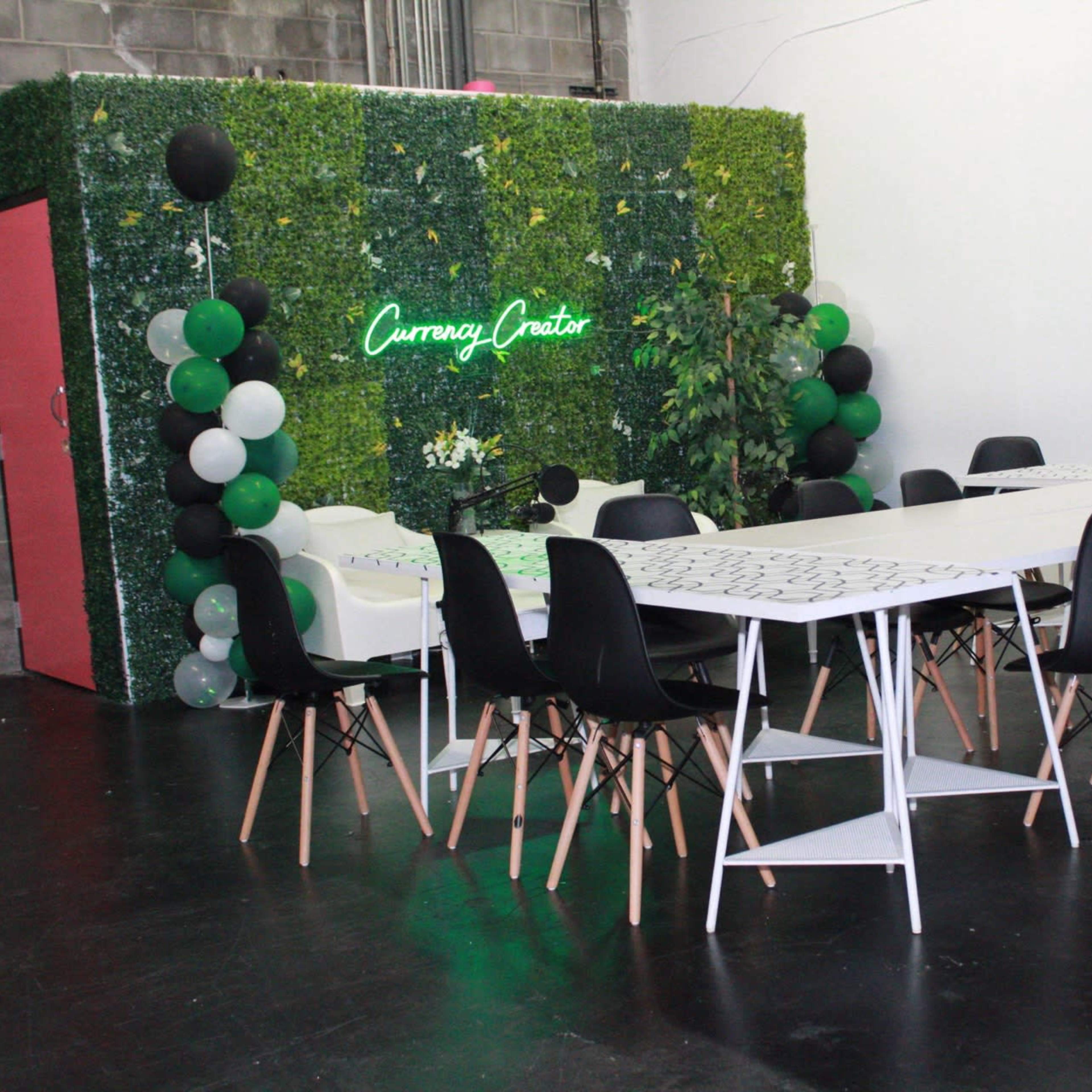 A large, well-lit meeting space features a long white table surrounded by black chairs, with a green decorative wall and a neon sign in the background.