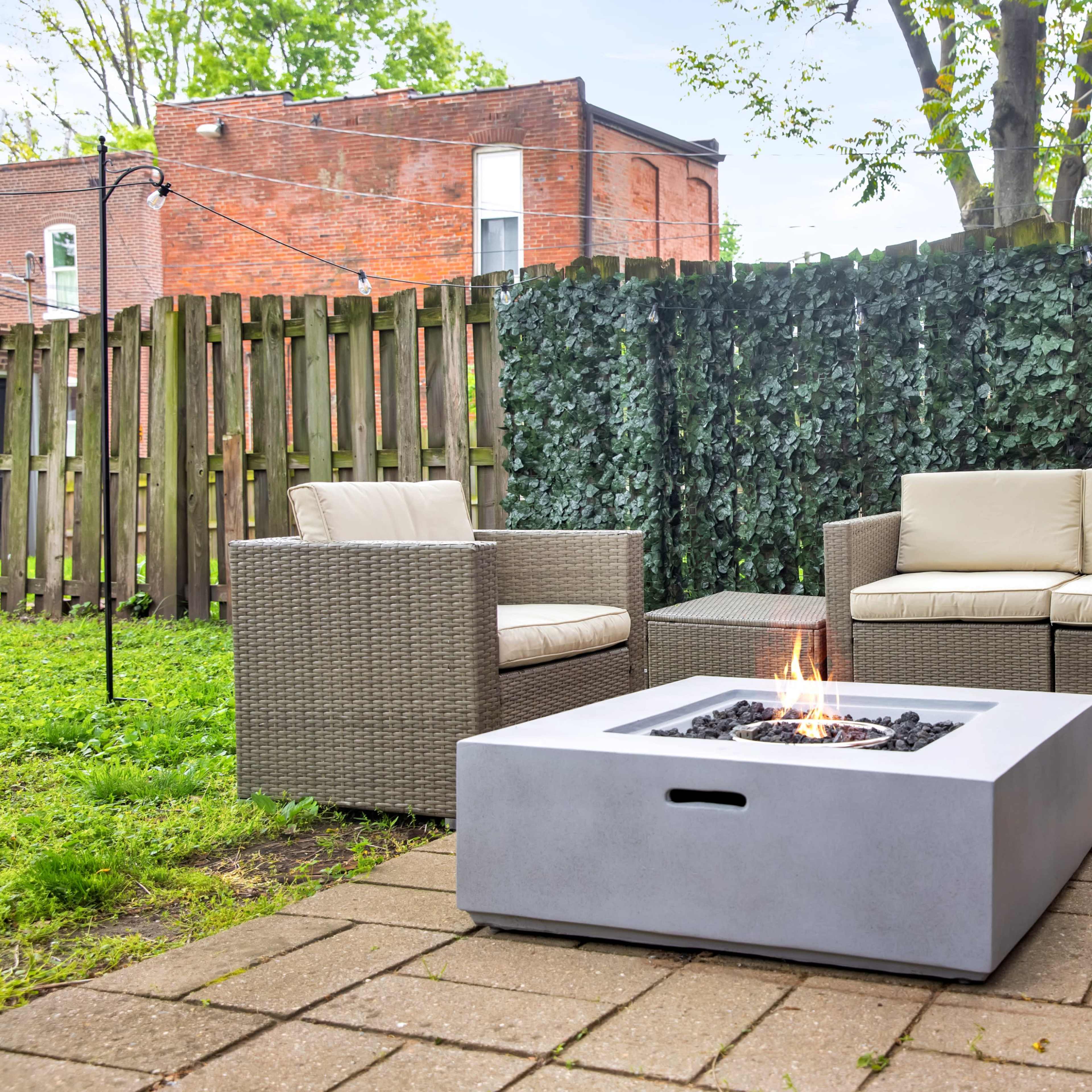 A modern outdoor seating area features a white fire pit surrounded by a sectional sofa and two armchairs on a stone patio, set against a backdrop of greenery and a wooden fence.