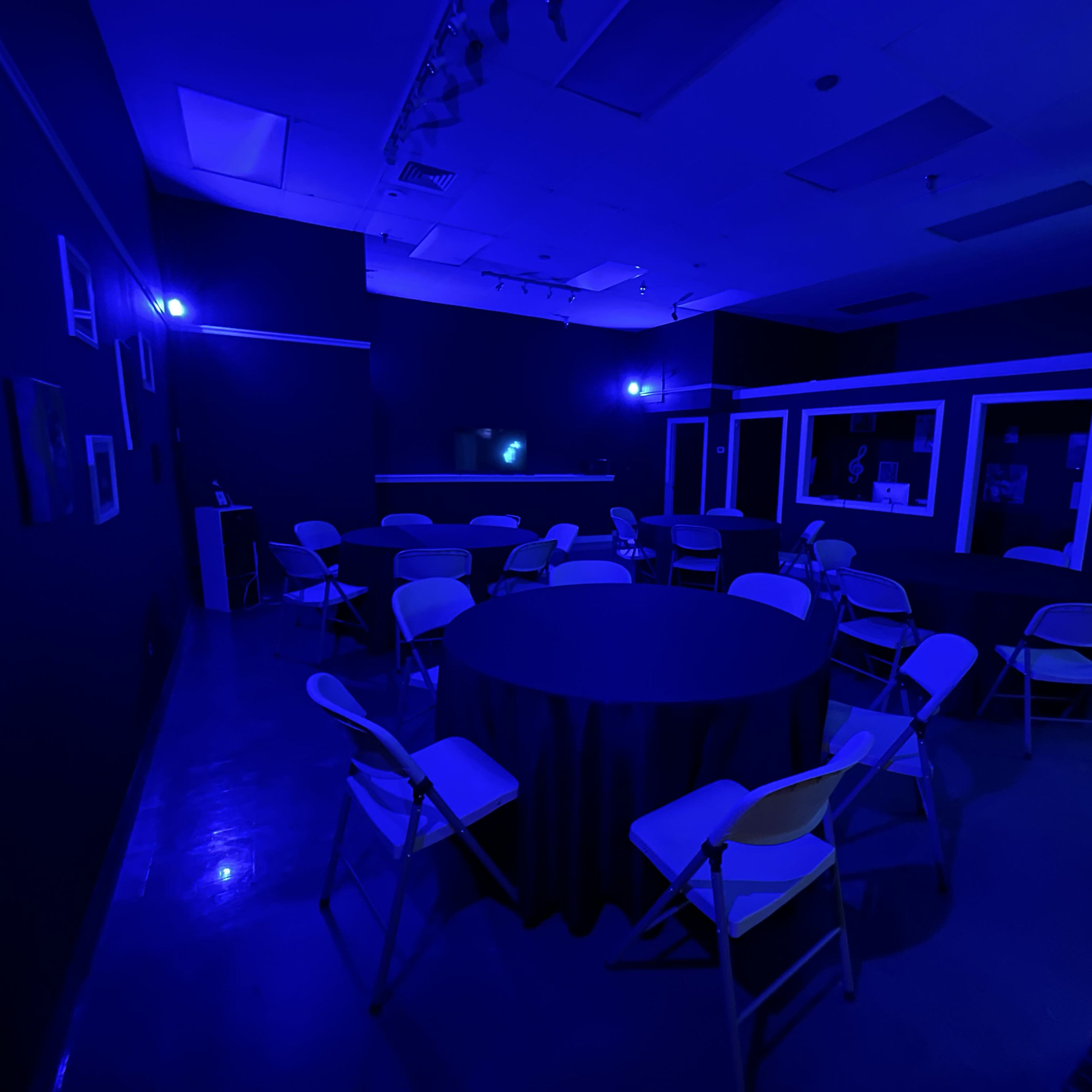 A dimly lit room with blue lighting, featuring a round table surrounded by folding chairs and a bar area in the background.