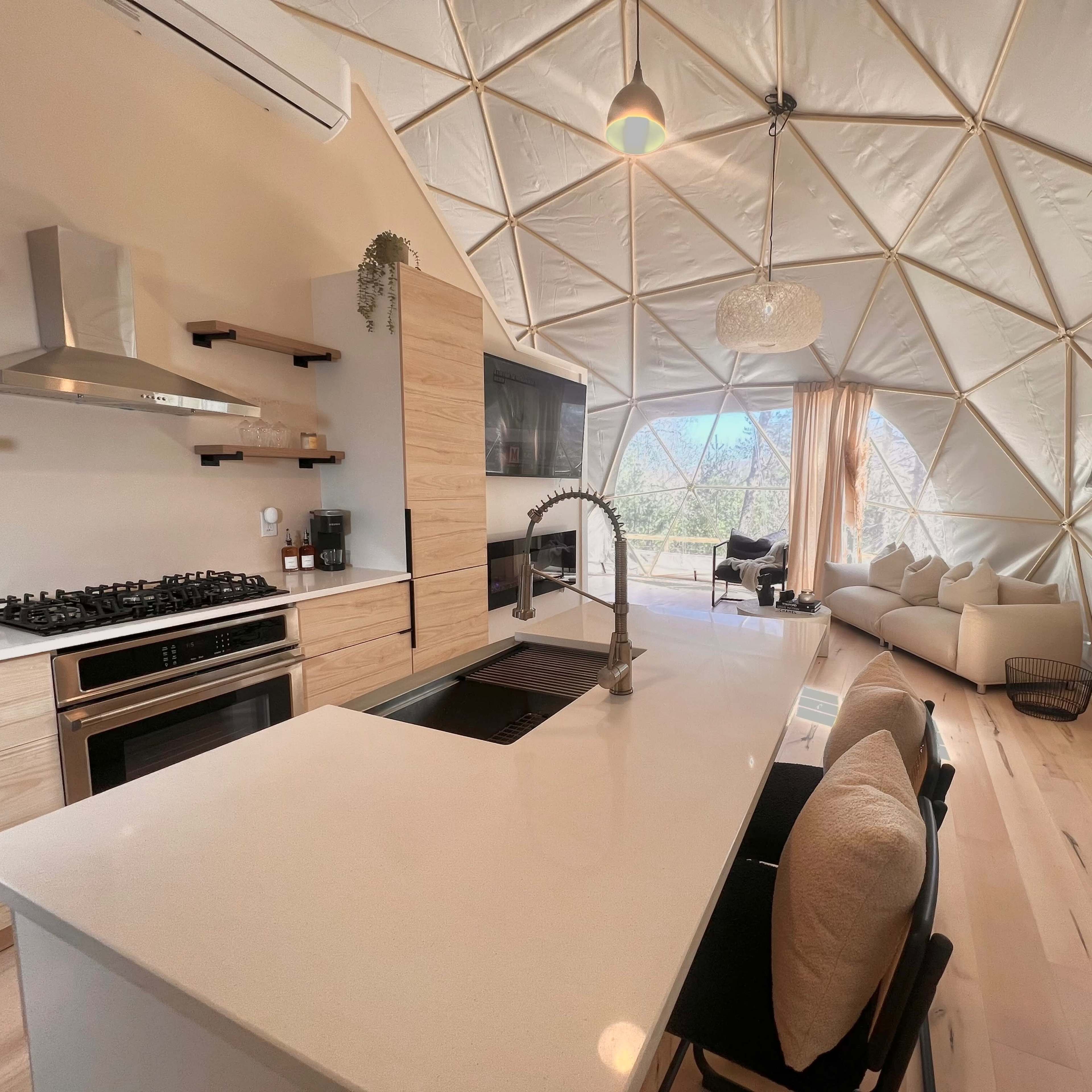 A modern kitchen and living area within a geodesic dome featuring light-colored wood cabinetry, a large island with a sink, and a cozy seating area with a view through large windows.