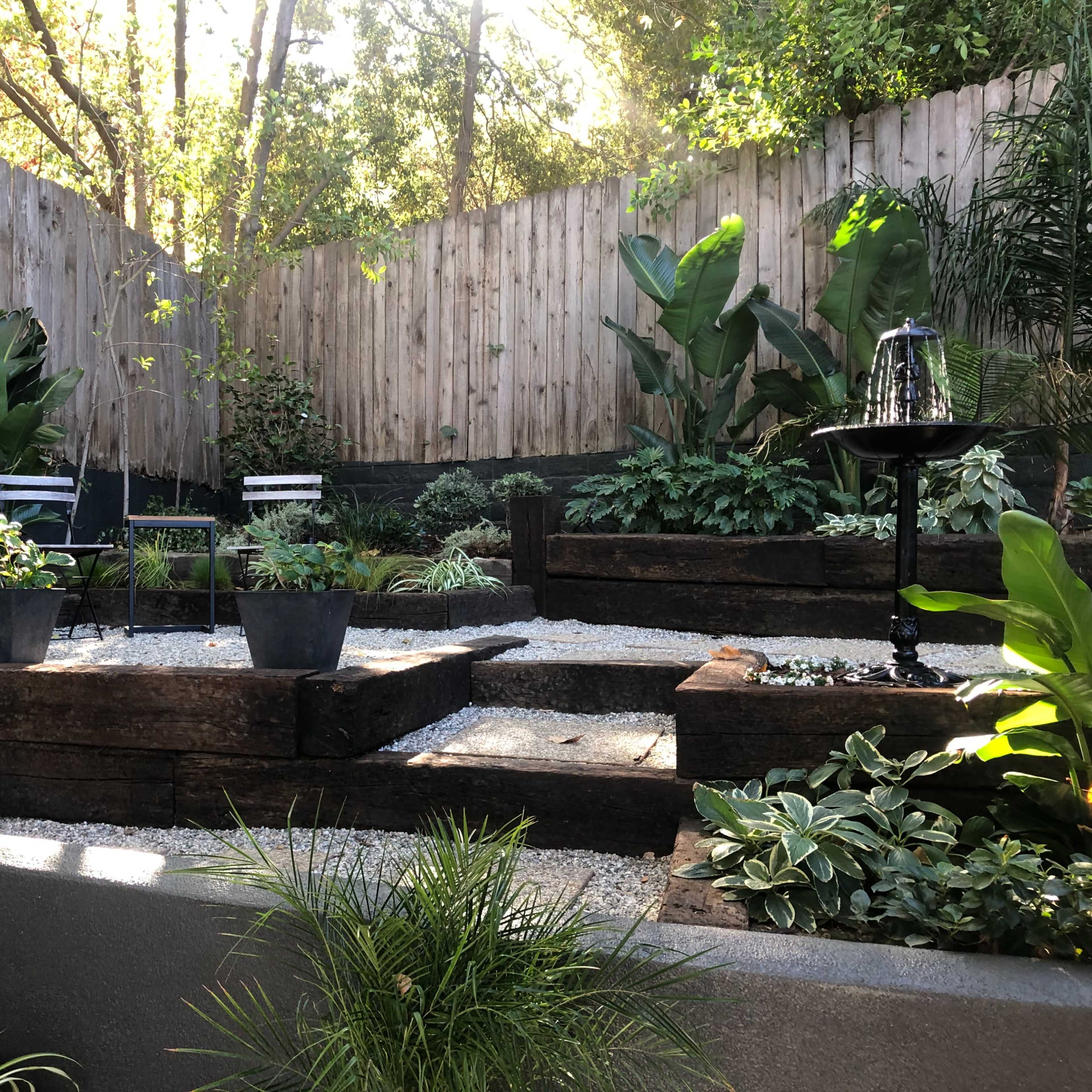 A landscaped garden with multiple levels, featuring potted plants, gravel paths, and wooden planters surrounded by greenery and a wooden fence.