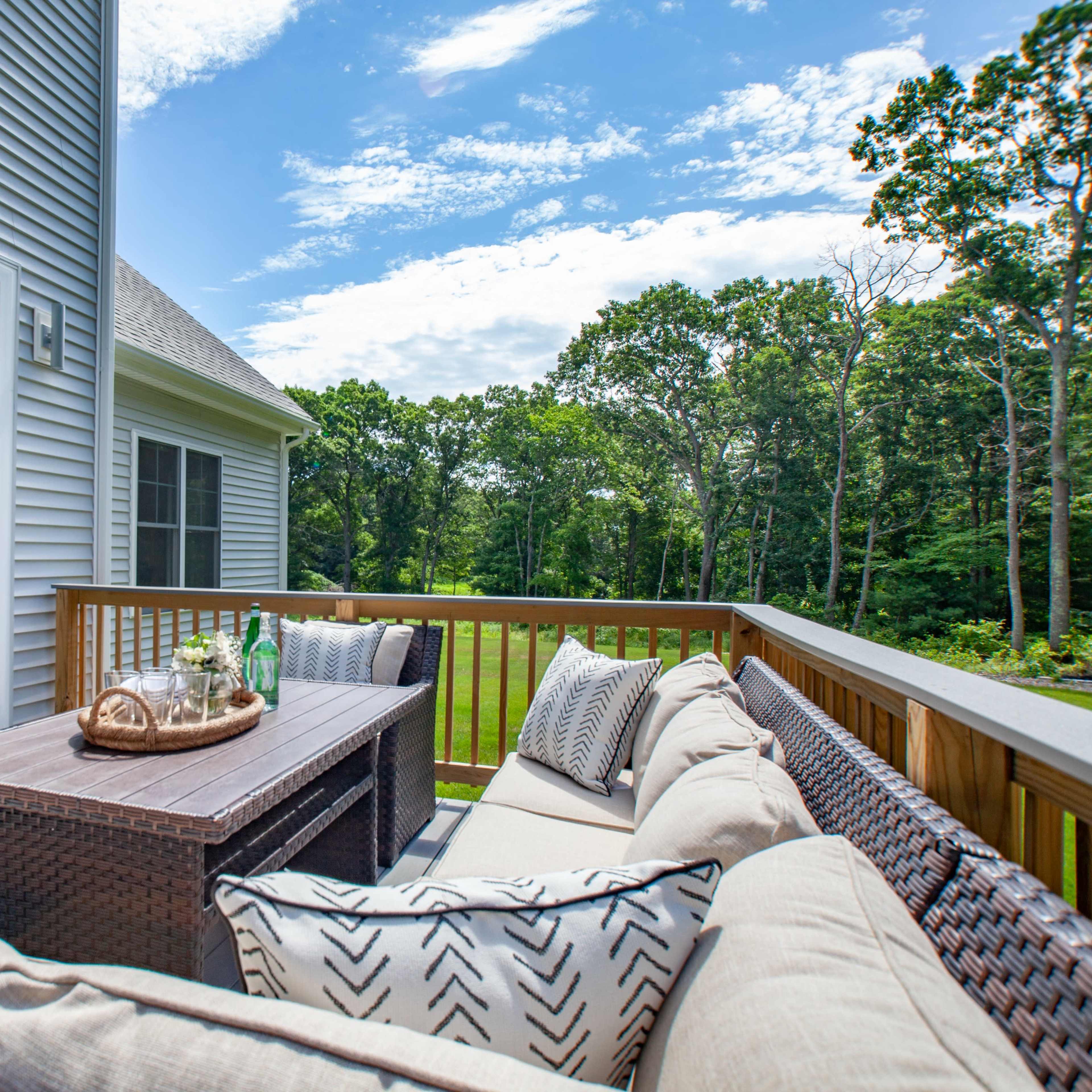 A patio area features a wicker seating arrangement with decorative cushions, a wooden table, and is surrounded by greenery under a partly cloudy sky.