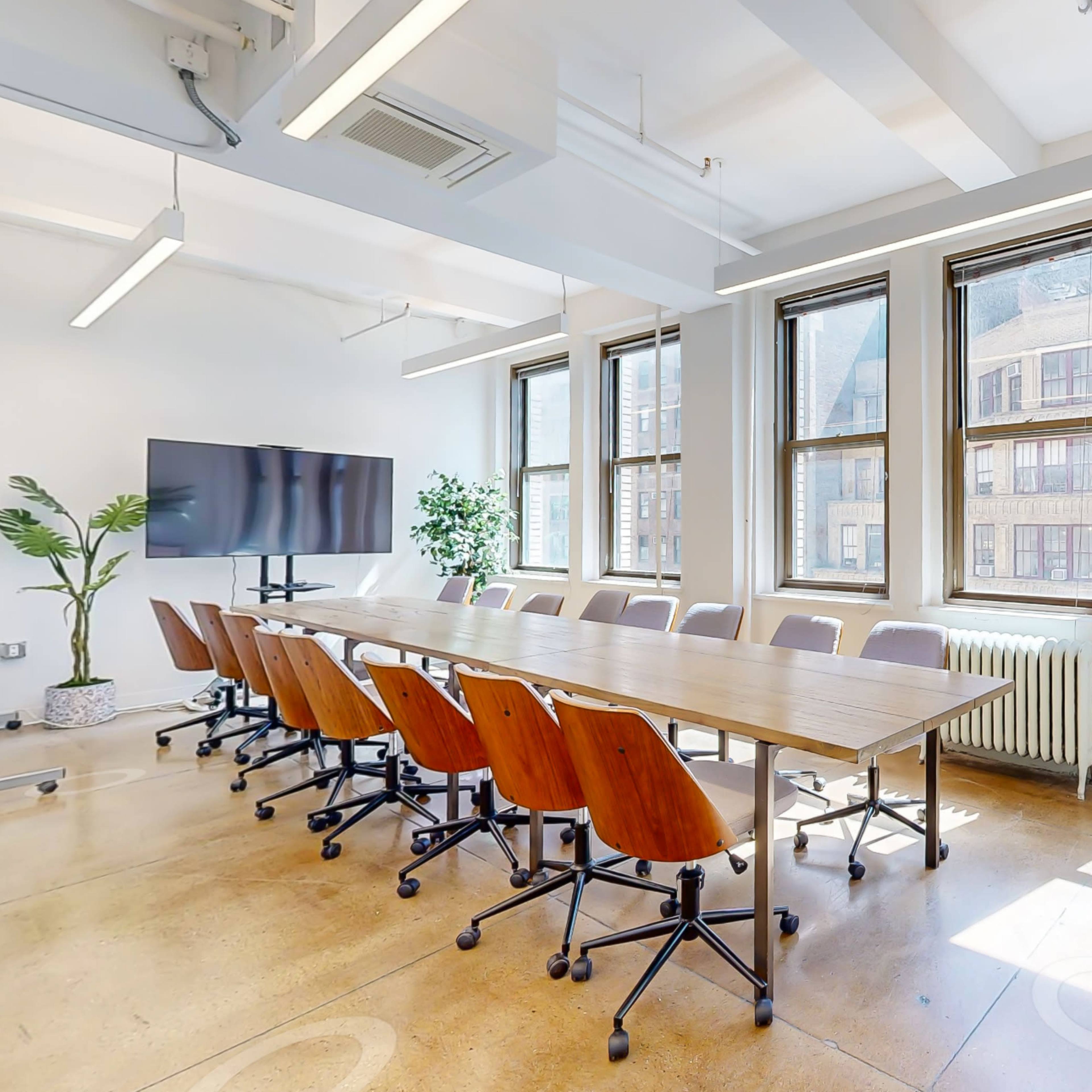 A spacious conference room features a long wooden table surrounded by ergonomic chairs, large windows letting in natural light, and plants adding greenery to the space.