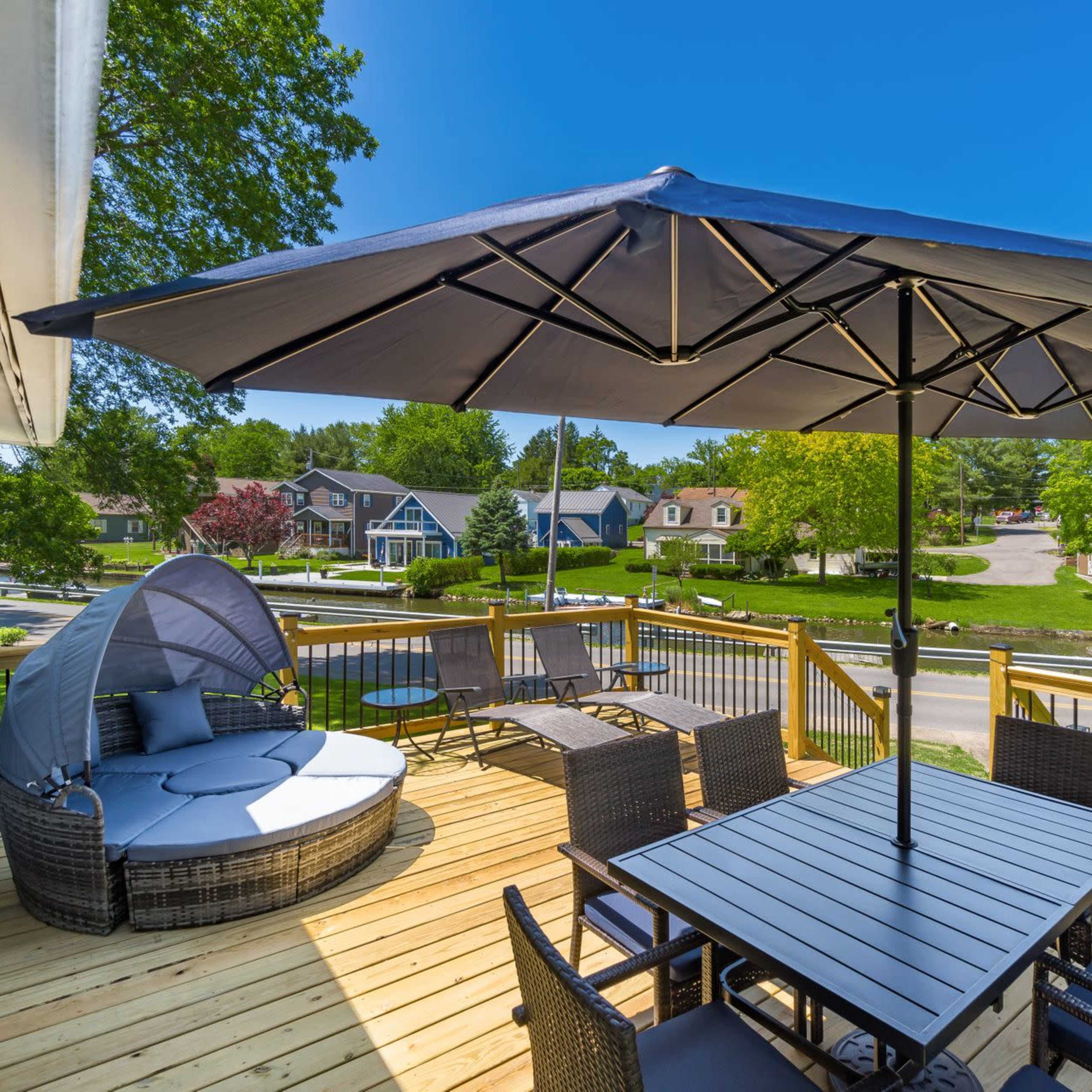 A wooden deck features a circular seating area with a large umbrella, a dining table set, and a view of nearby houses and greenery.