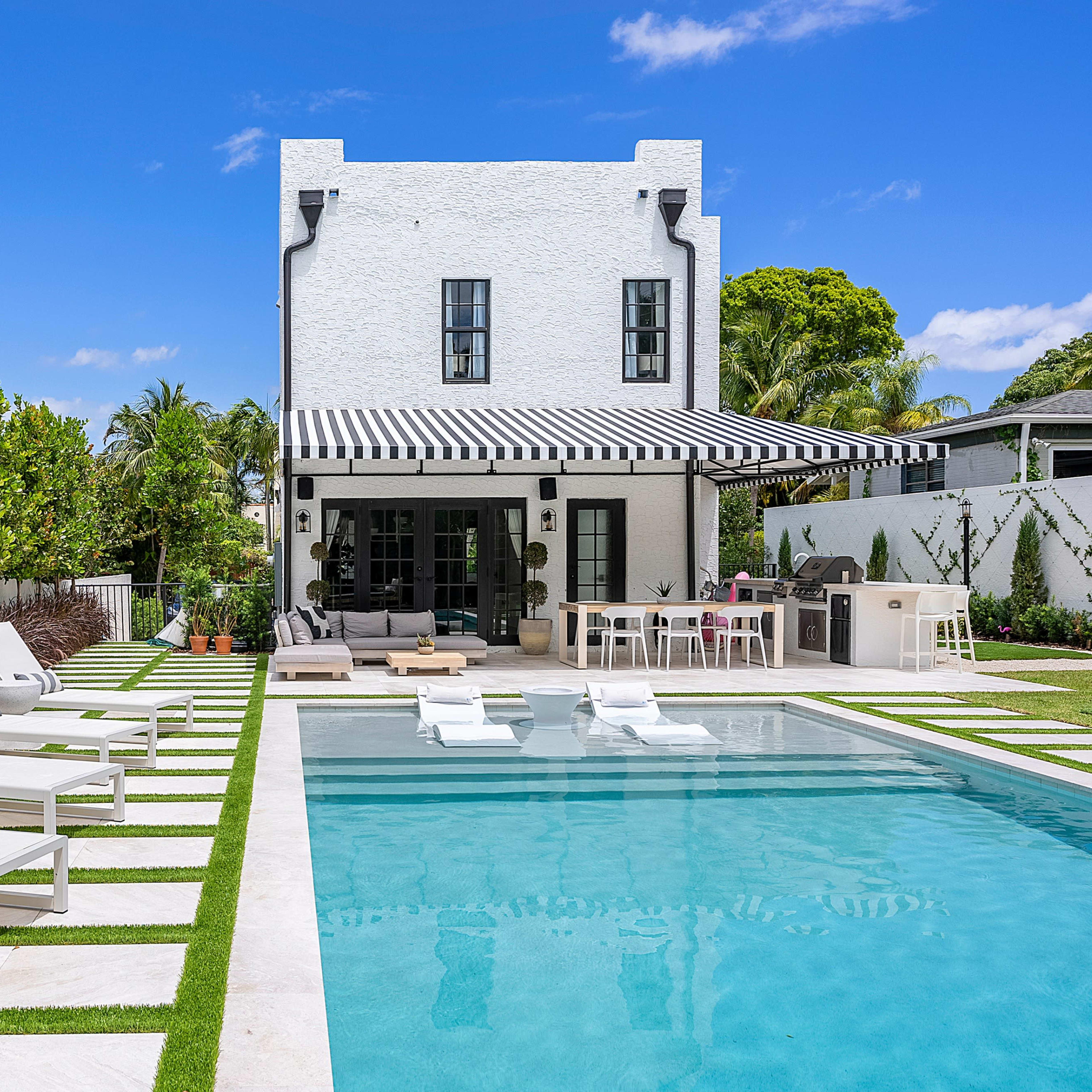A modern backyard with a swimming pool, lounge chairs, and a two-story white house surrounded by greenery.