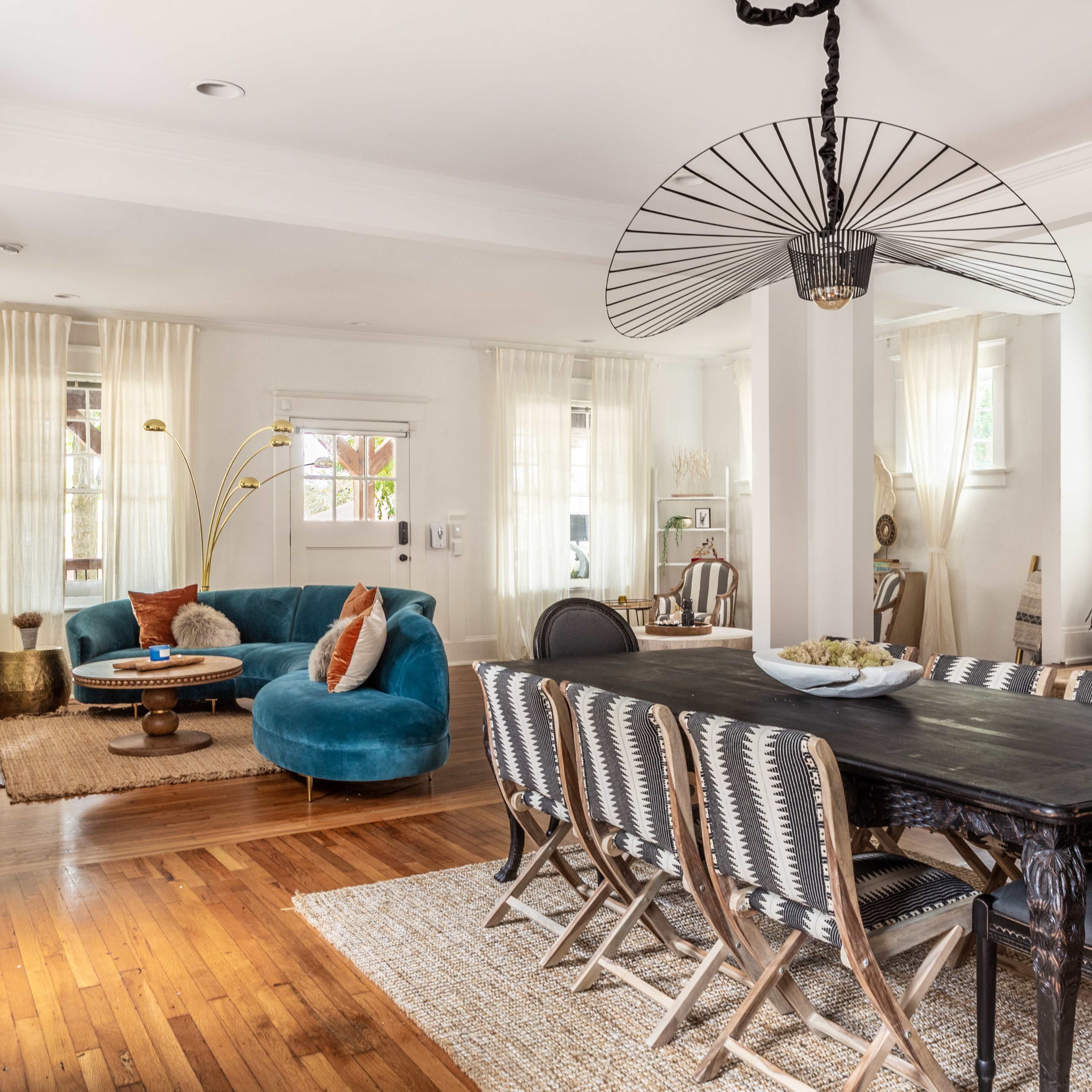 A spacious living and dining area with a turquoise sofa, a long dark wooden dining table, and bright curtains allowing natural light to illuminate the room.