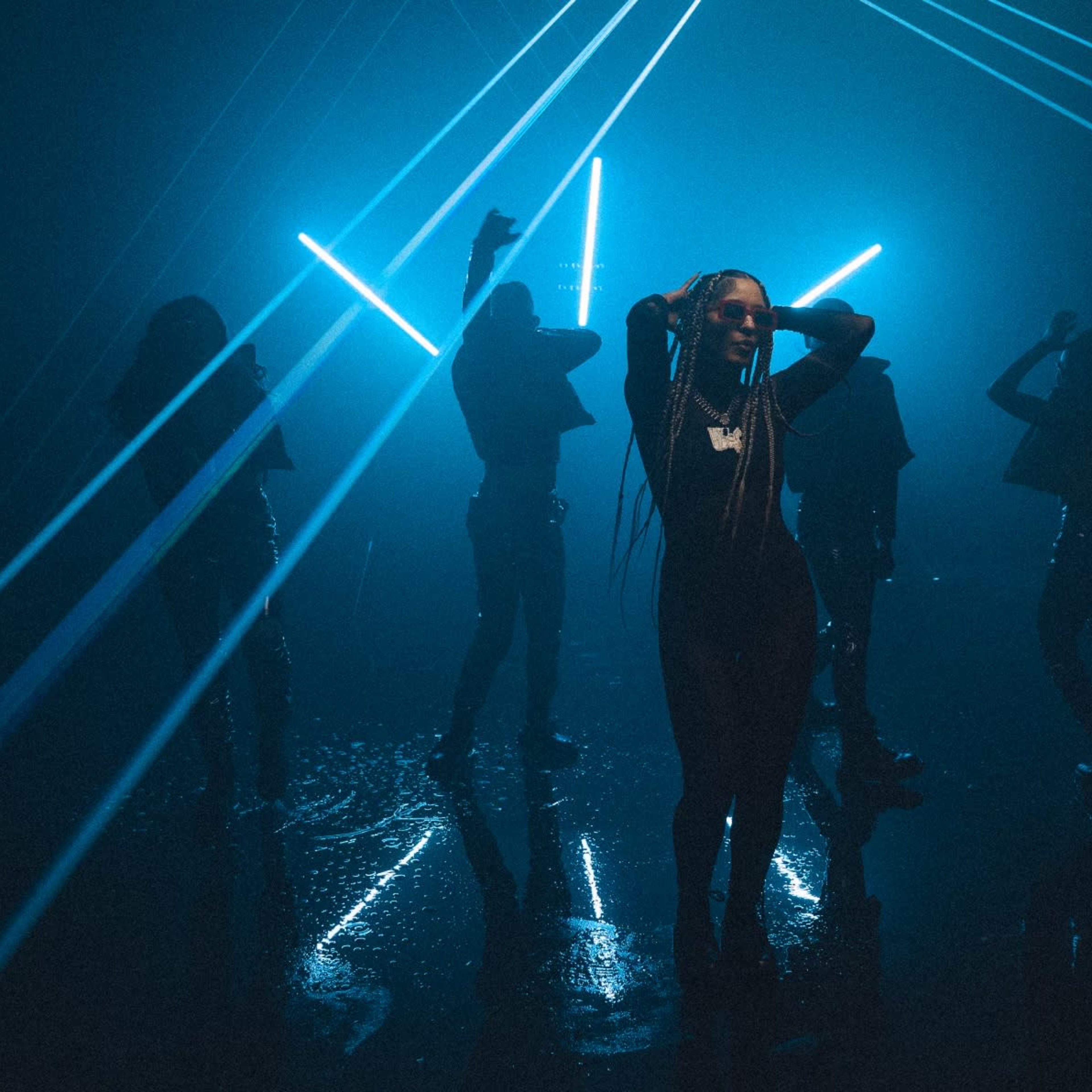 A group of silhouetted dancers illuminated by bright blue laser beams in a dimly lit space.
