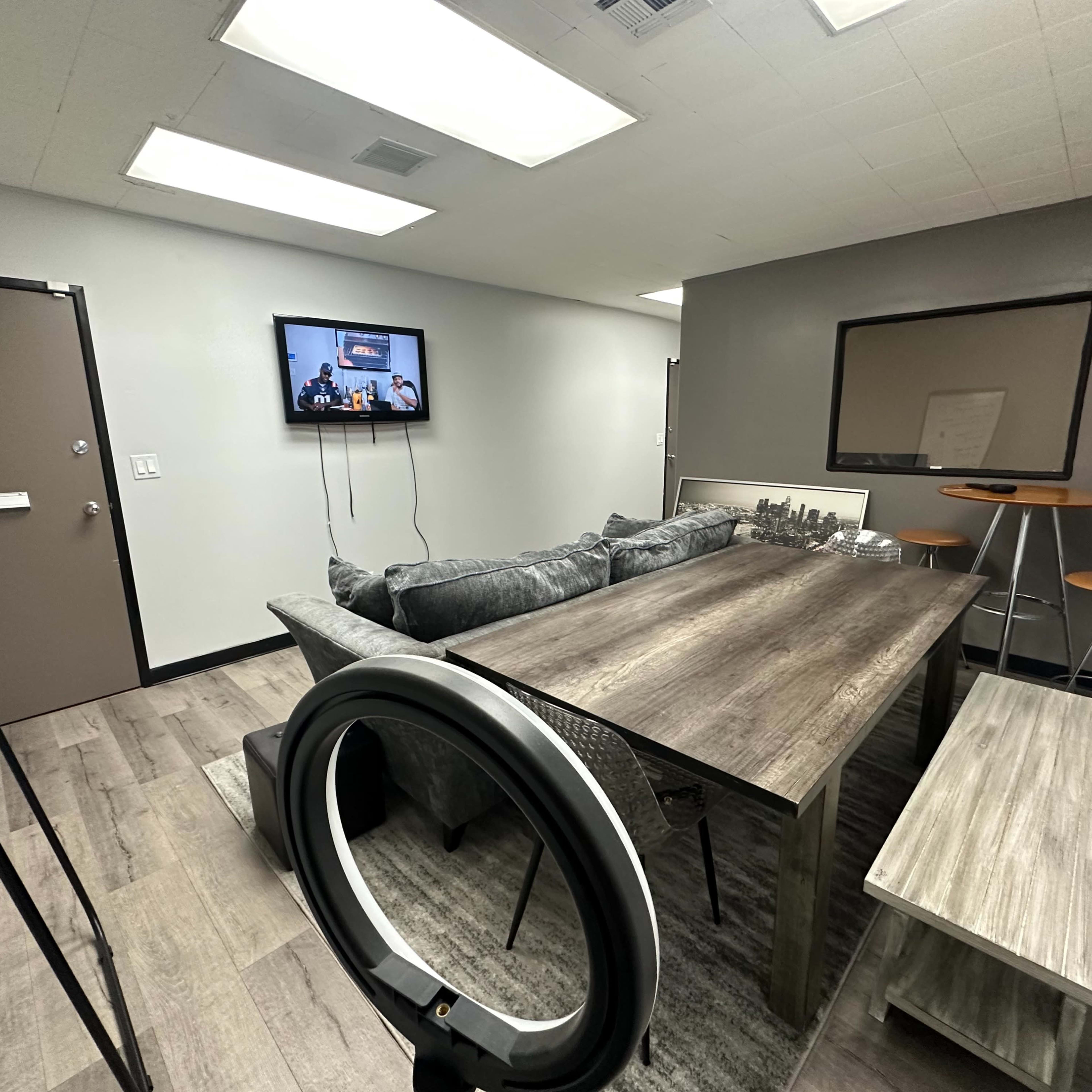 A modern conference room equipped with a wooden table, a large TV on the wall, a mirror, and seating arrangements including a sofa and a stool.
