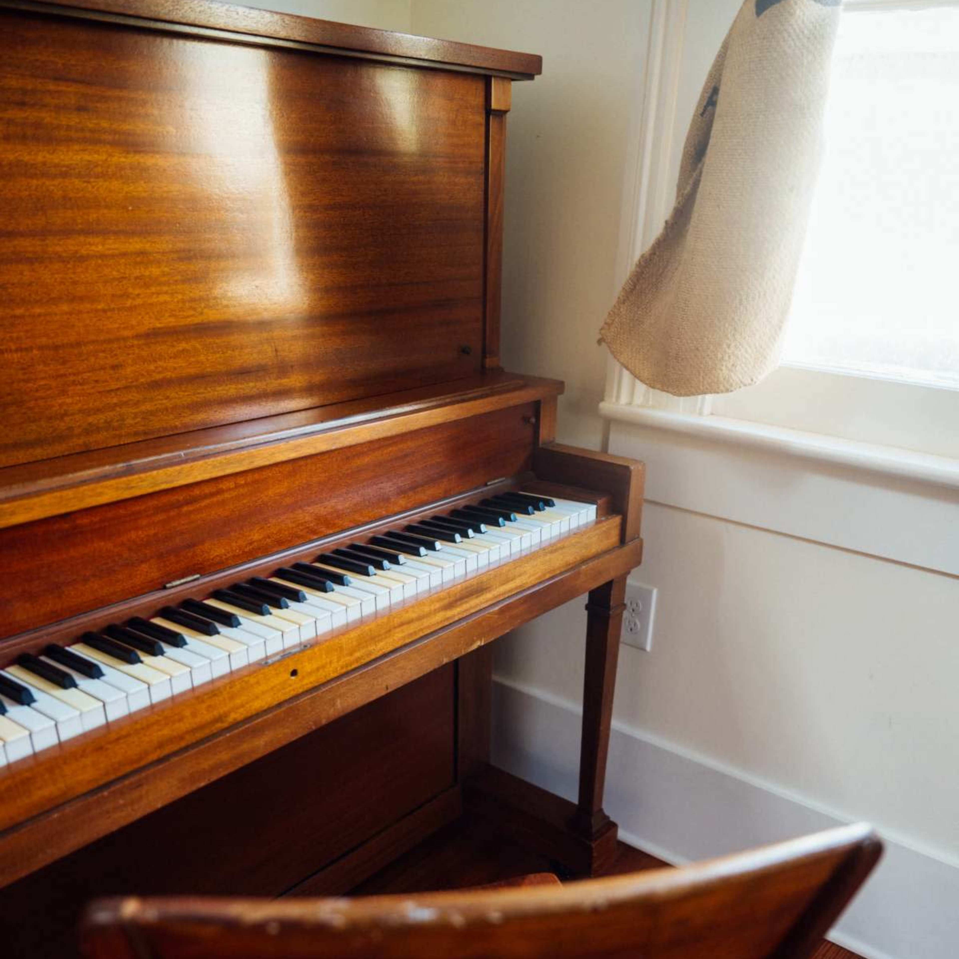 A wooden piano with a row of black and white keys is positioned next to a window, complemented by a wooden chair facing it.