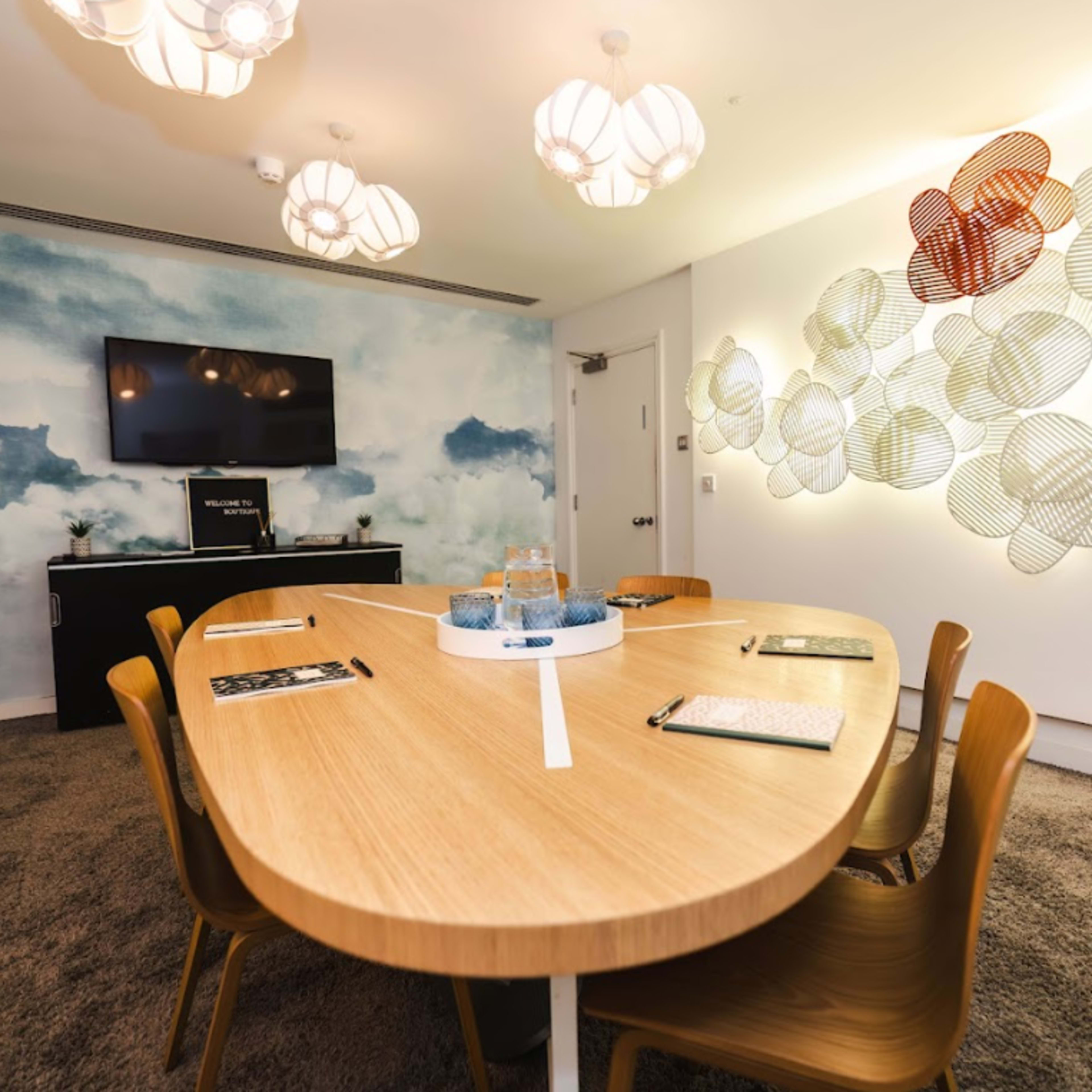 A modern conference room with a long wooden table, chairs, a TV mounted on the wall, and a decorative mural featuring clouds.