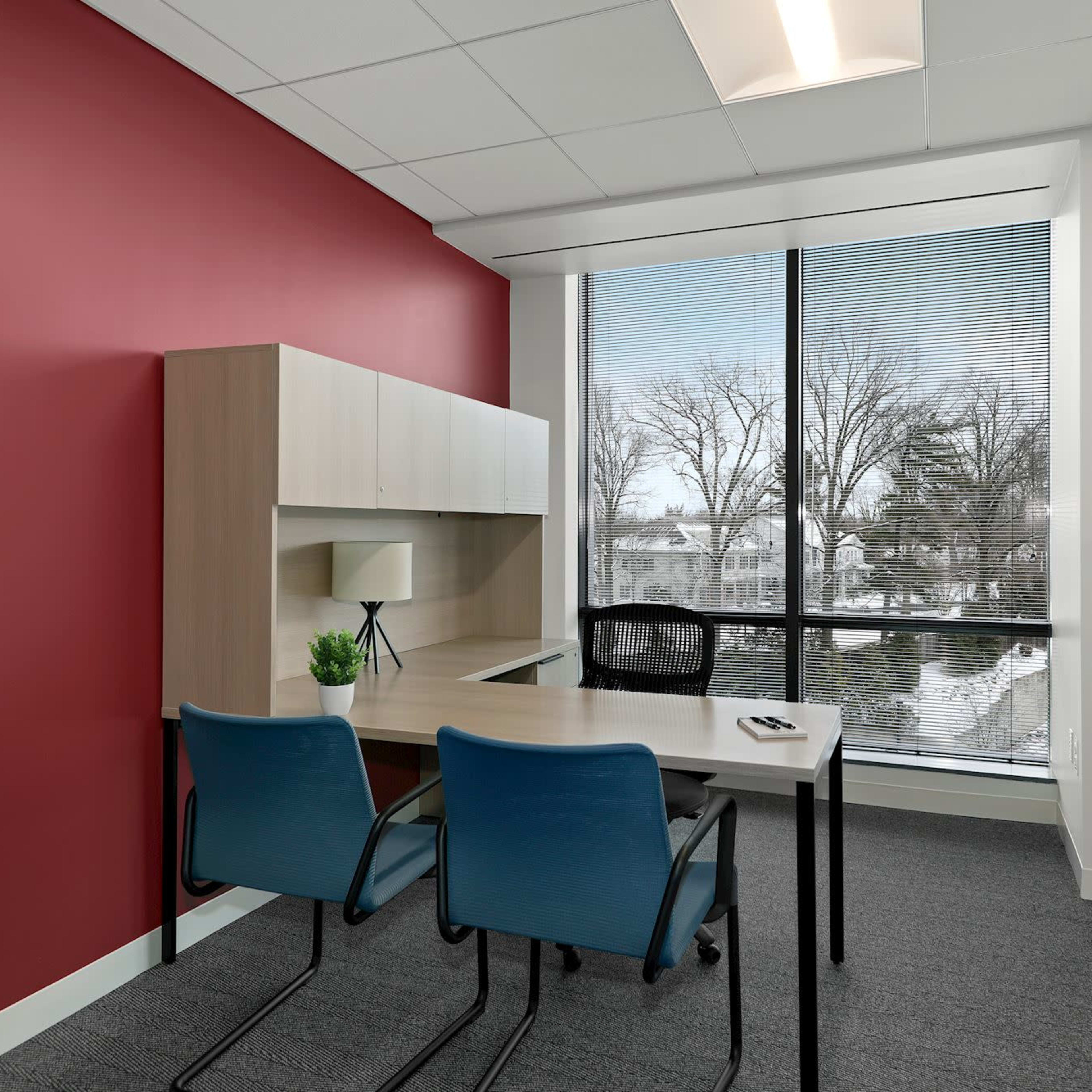 A small office space featuring a desk with two blue chairs, a light-colored office cabinet, and a large window overlooking a snowy landscape.